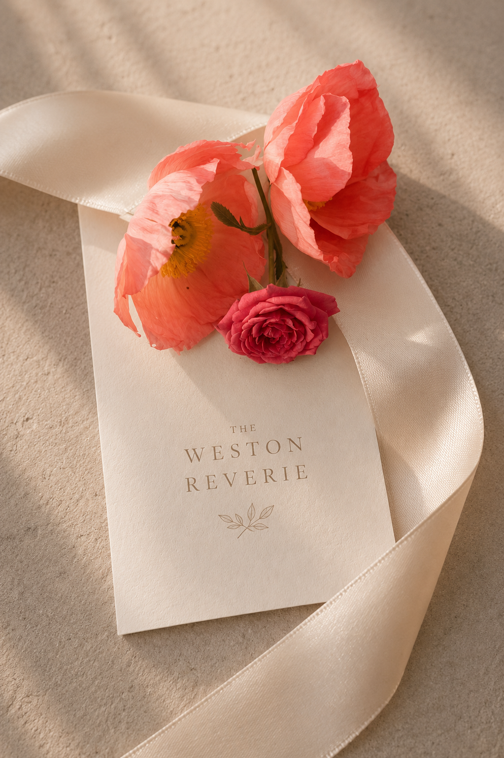 A floral arrangement with pink poppies and a red rose resting on an invitation card that reads 'The Weston Reverie,' adorned with a satin ribbon on a soft, neutral surface.