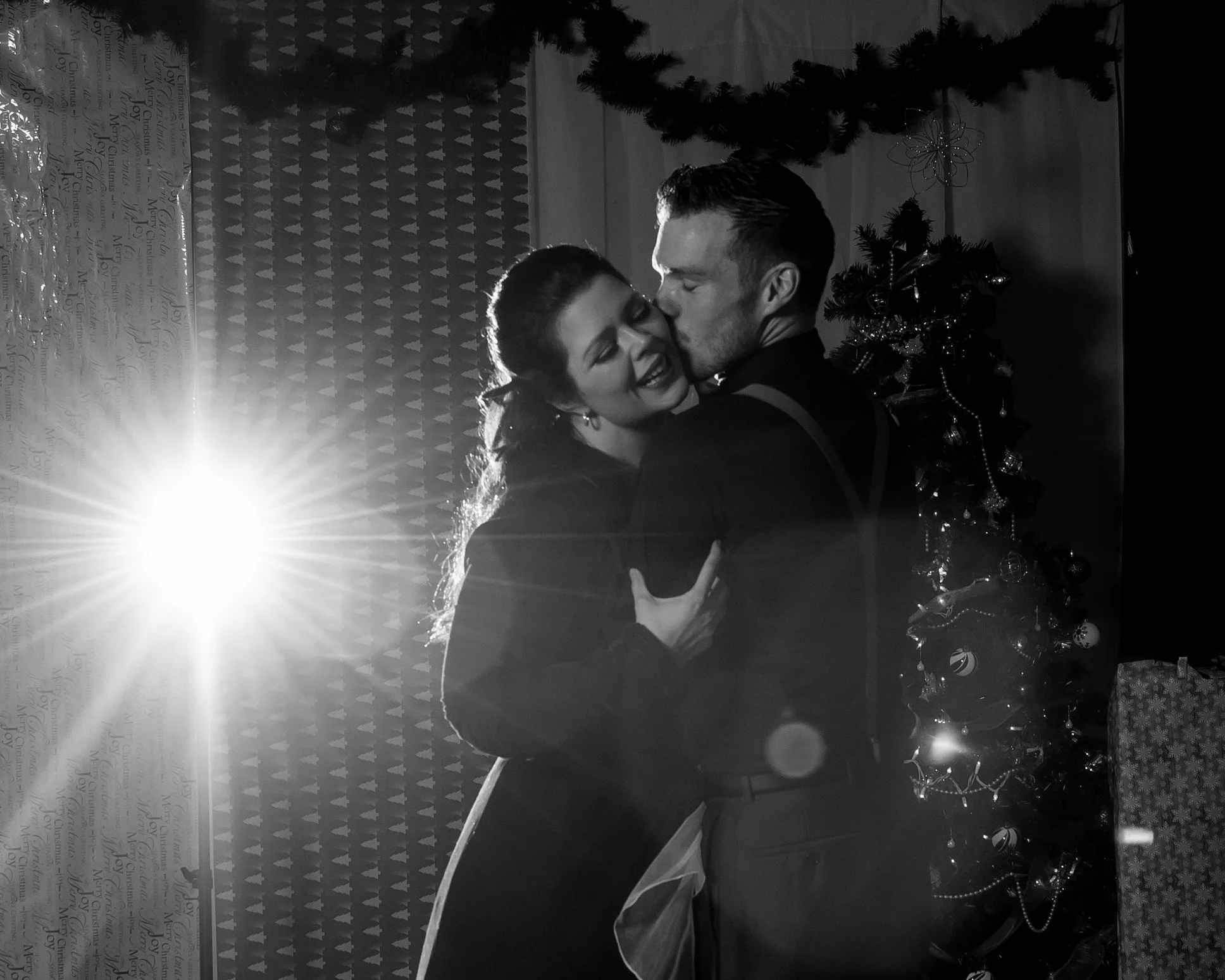 A black and white photo of a couple dancing in front of a Christmas tree, with holiday decorations and wrapped gifts visible in the background, lit by a bright light source on the left side.
