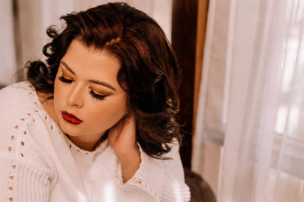 A woman with dark wavy hair and red lipstick is resting her head on her hand, looking down and to the side, sitting near a window with sheer curtains.