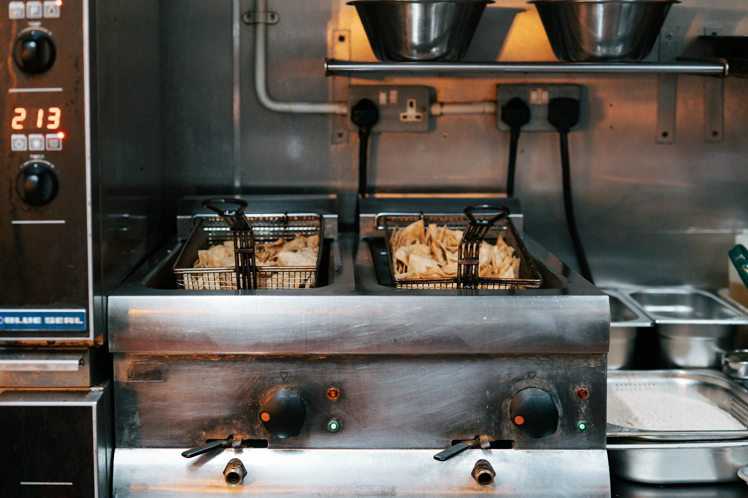 Commercial deep fryer with two baskets filled with fried food, likely potato chips or fries, in a professional kitchen setting.