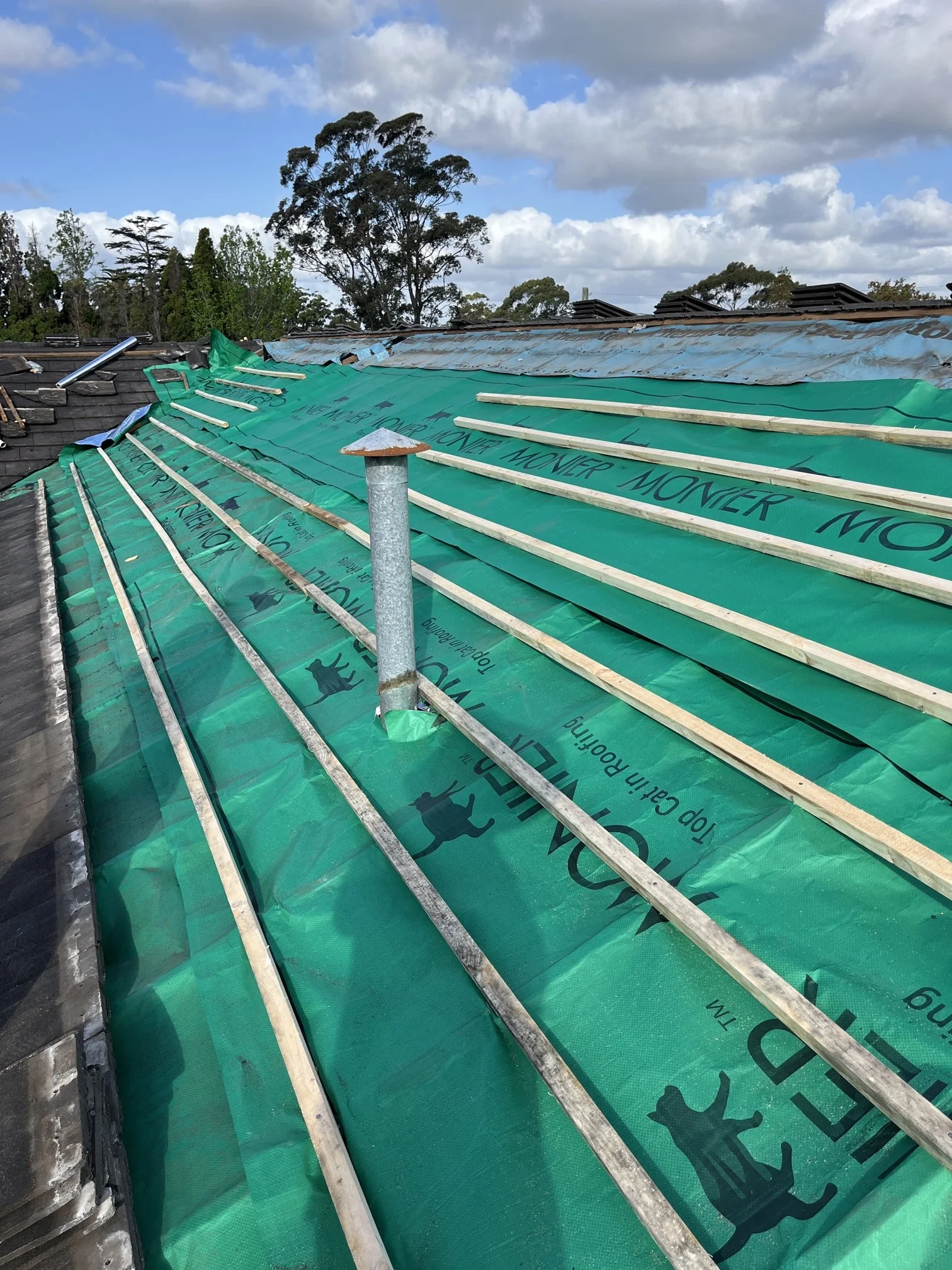 Roof under construction with green weatherproofing membrane, wooden battens, and some sections of slate tiles. The background shows a partly cloudy sky and trees.