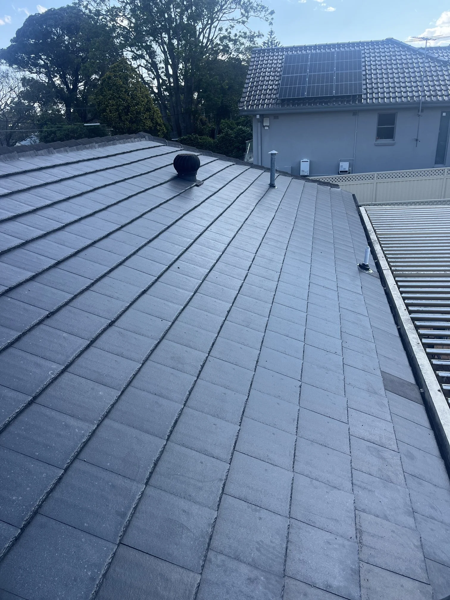 View of a roof with solar panels on a residential house, with trees in the background and a partly cloudy sky.