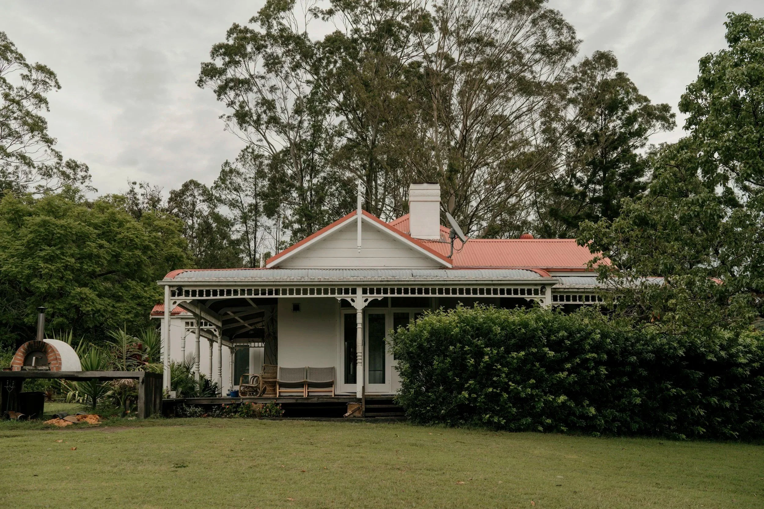 A white house with a red roof, chimney, and a small porch, surrounded by trees and shrubs on a cloudy day.