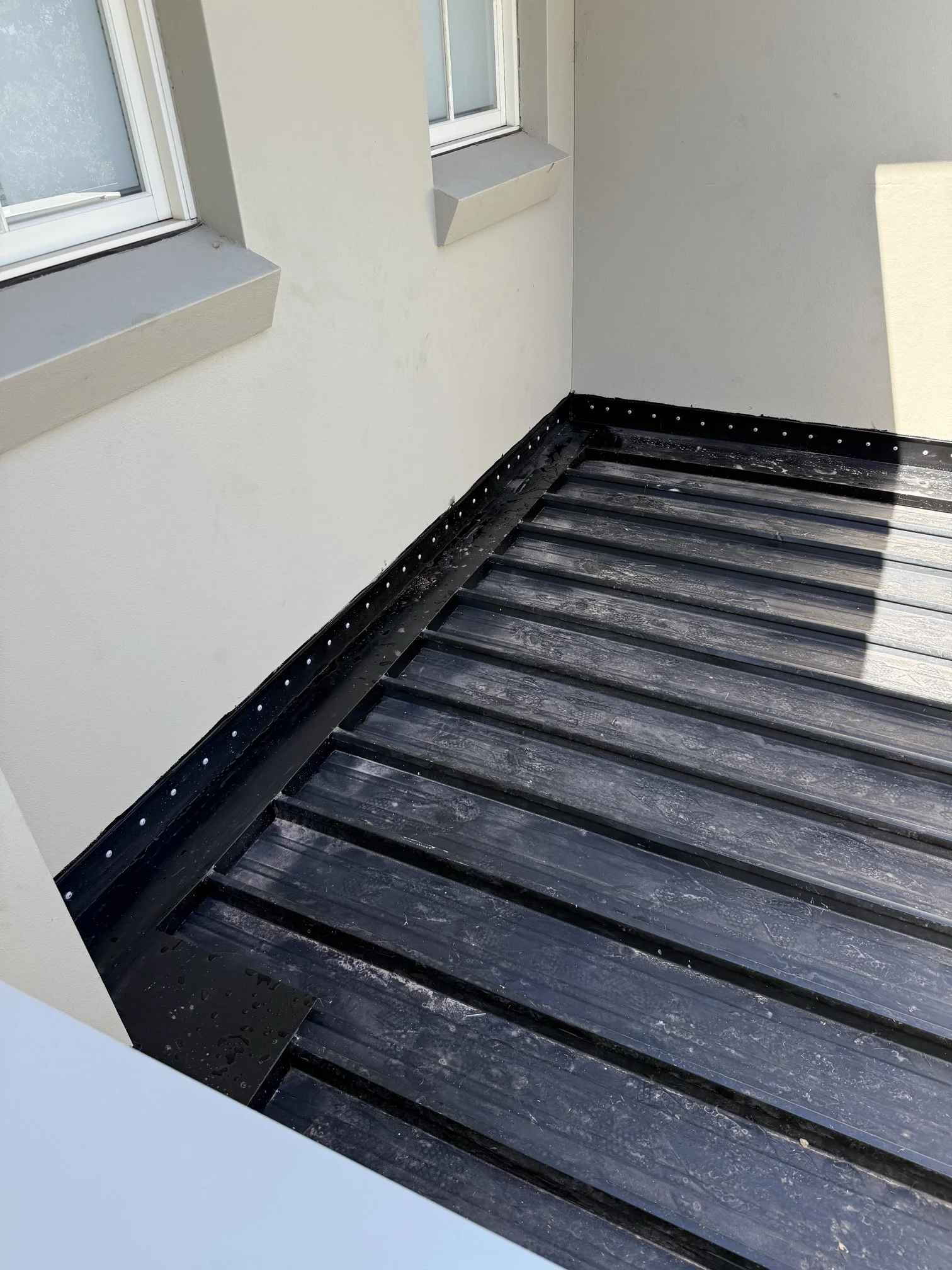 Close-up of a balcony with black wooden decking and a white wall with two windows. The decking has some water droplets on it.