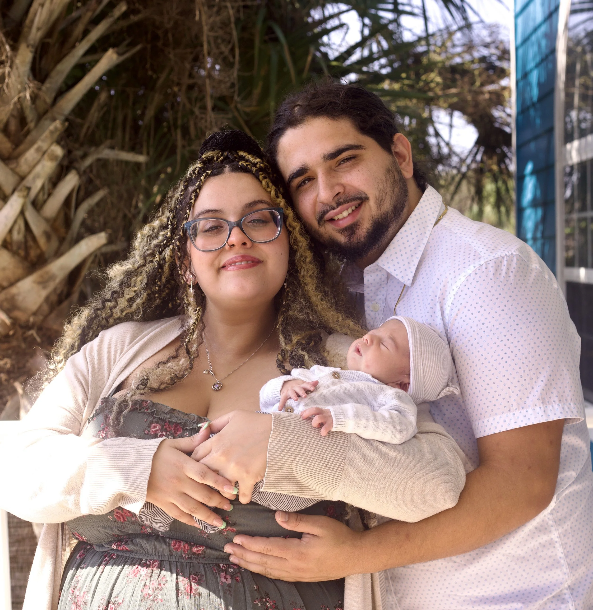 A diverse group of people, including a woman with glasses and a man, holding a baby, standing outdoors with trees and a building in the background.
