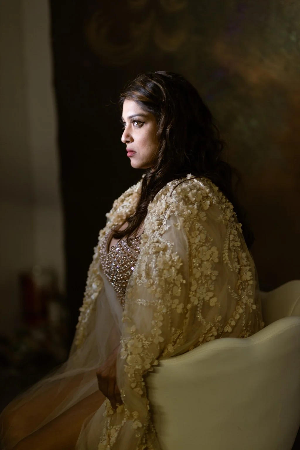 A woman with dark wavy hair sitting in profile, wearing a beige dress with embellishments.
