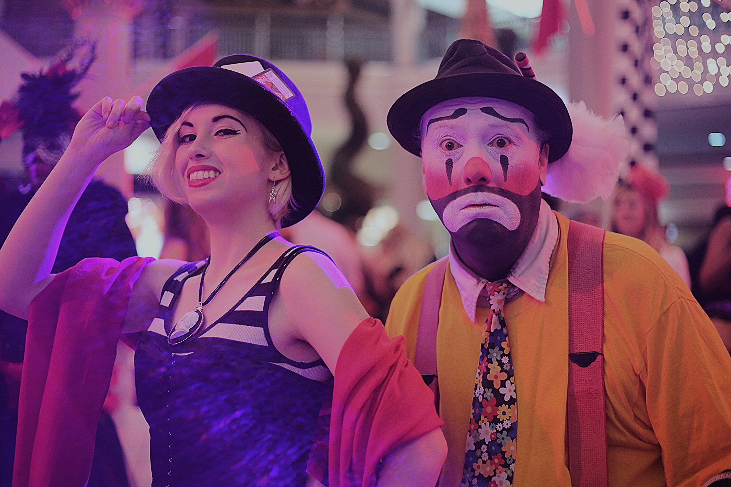 Young woman with blonde hair, face painted as a witch, wearing a witch hat and a black and white striped top, smiling and looking at the camera; man dressed as a clown with face paint, orange shirt, suspenders, and a brown hat, standing next to her in an amusement park or carnival setting with colorful lights.
