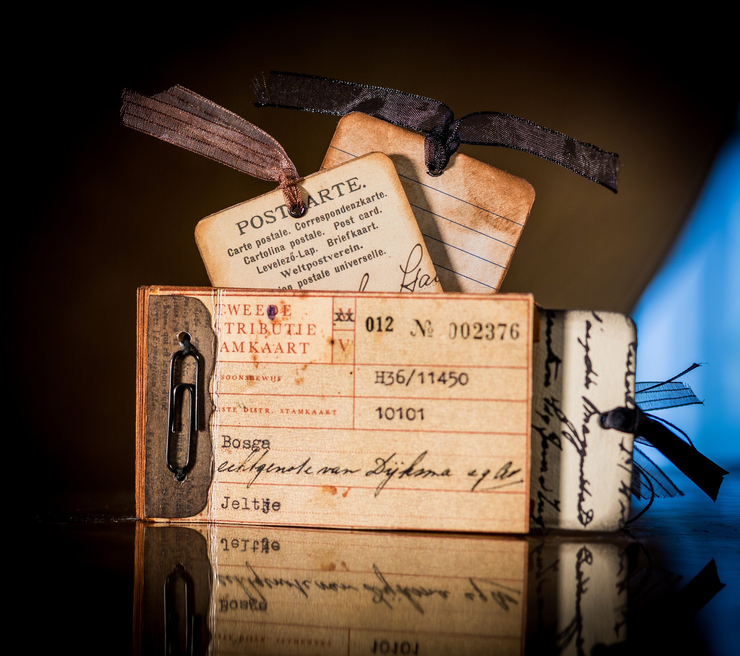 Close-up of vintage postcards, lined up with ribbons attached, against a dark background. The postcards are aged and feature handwritten text and printed details.