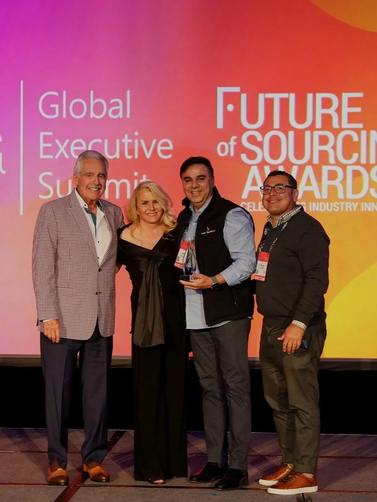 Four people standing on a stage at the Global Executive Summit, with a large pink and orange screen behind them displaying the text 'Future of Sourcing Awards.' One person is holding an award, and they are all smiling.