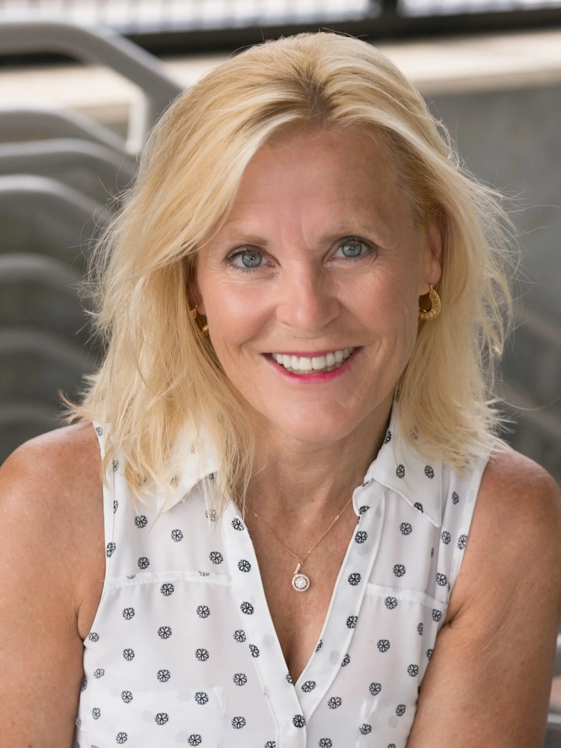 Close-up of a woman with blonde hair, blue eyes, and a smile, wearing a white sleeveless top with black floral patterns, gold earrings, and a pendant necklace, with gray chairs in the background.