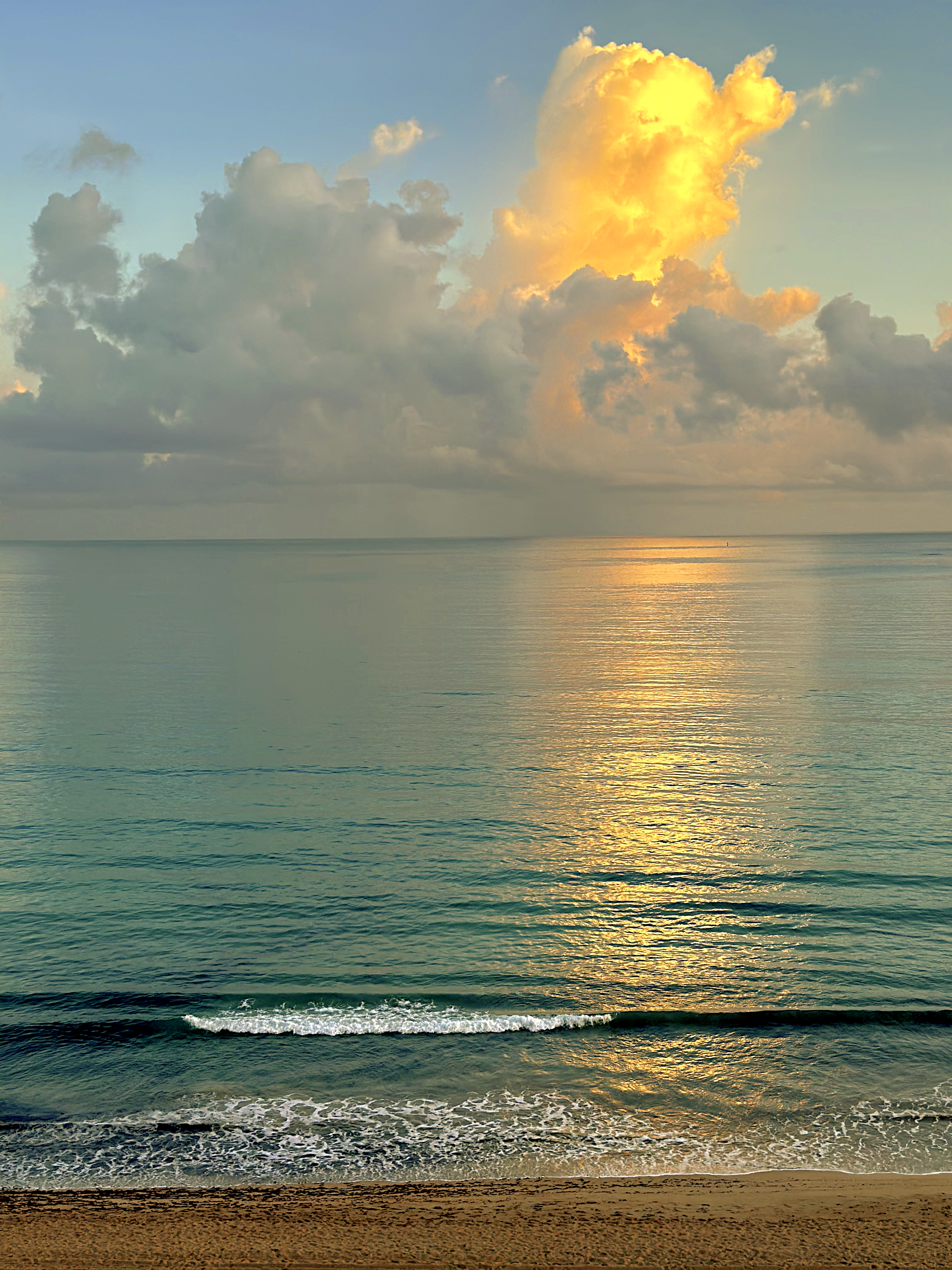 A serene view of the ocean at sunset with golden clouds reflected on the calm water, near a sandy beach.