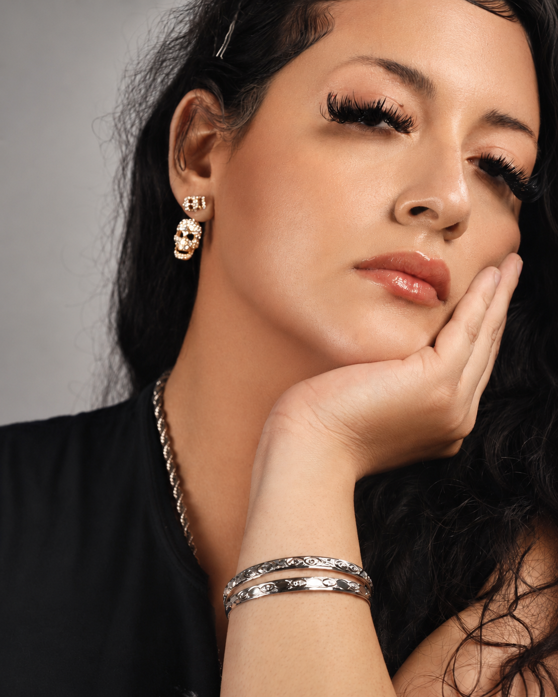 Close-up of a woman with dark curly hair wearing skull-shaped earrings, a silver bracelet, and a black top, resting her face on her hand.