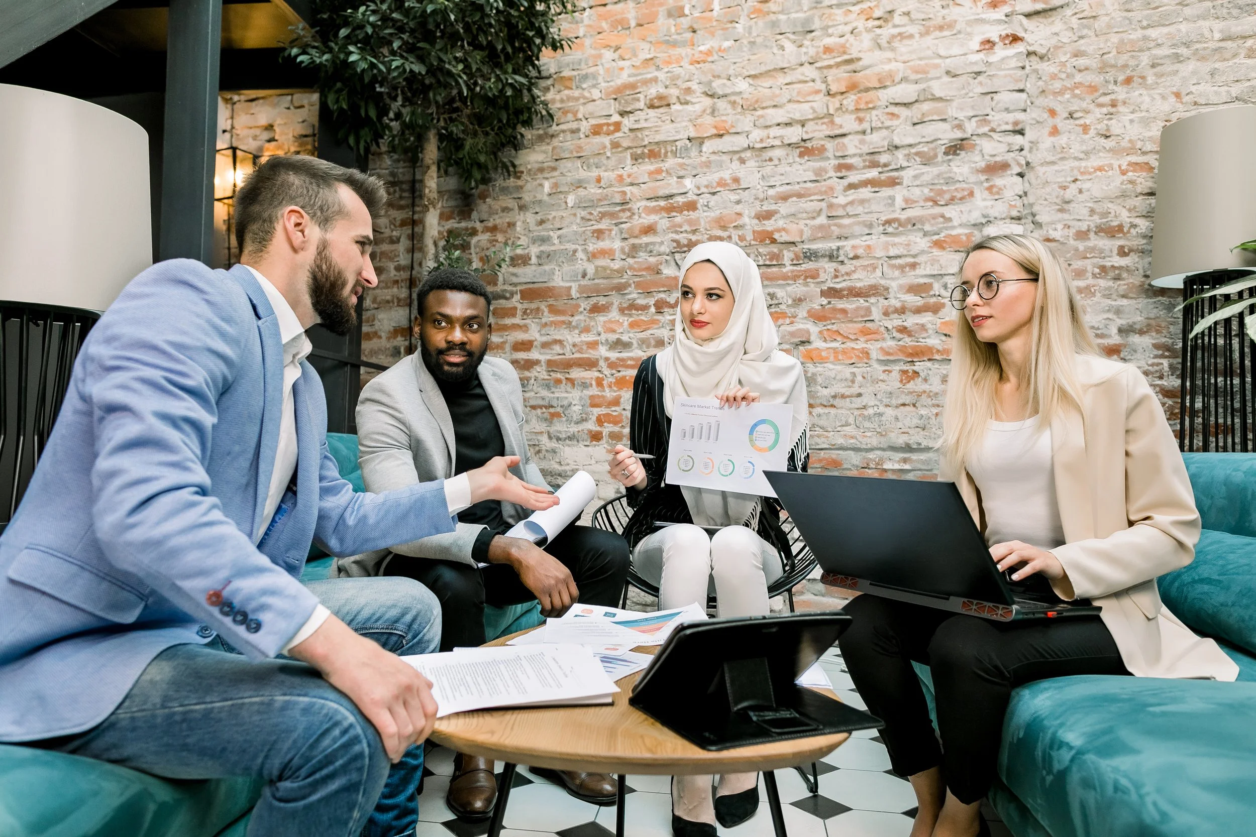 Group of five diverse professionals in business casual attire participating in a discussion or meeting in a modern office with exposed brick walls and decorative plants.