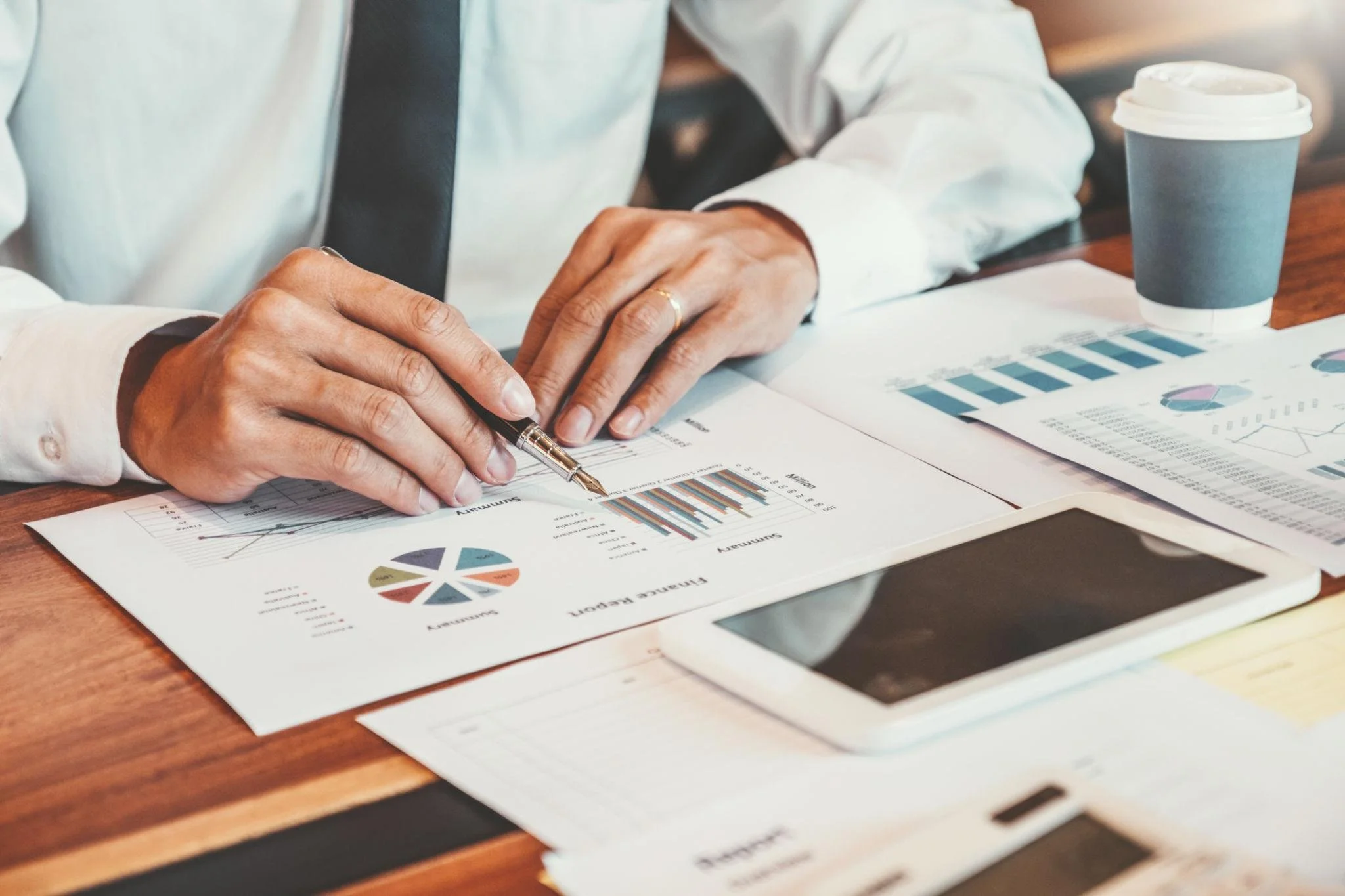 Close-up of a person reviewing financial documents with graphs and charts, sitting at a wooden desk, with a disposable coffee cup nearby.