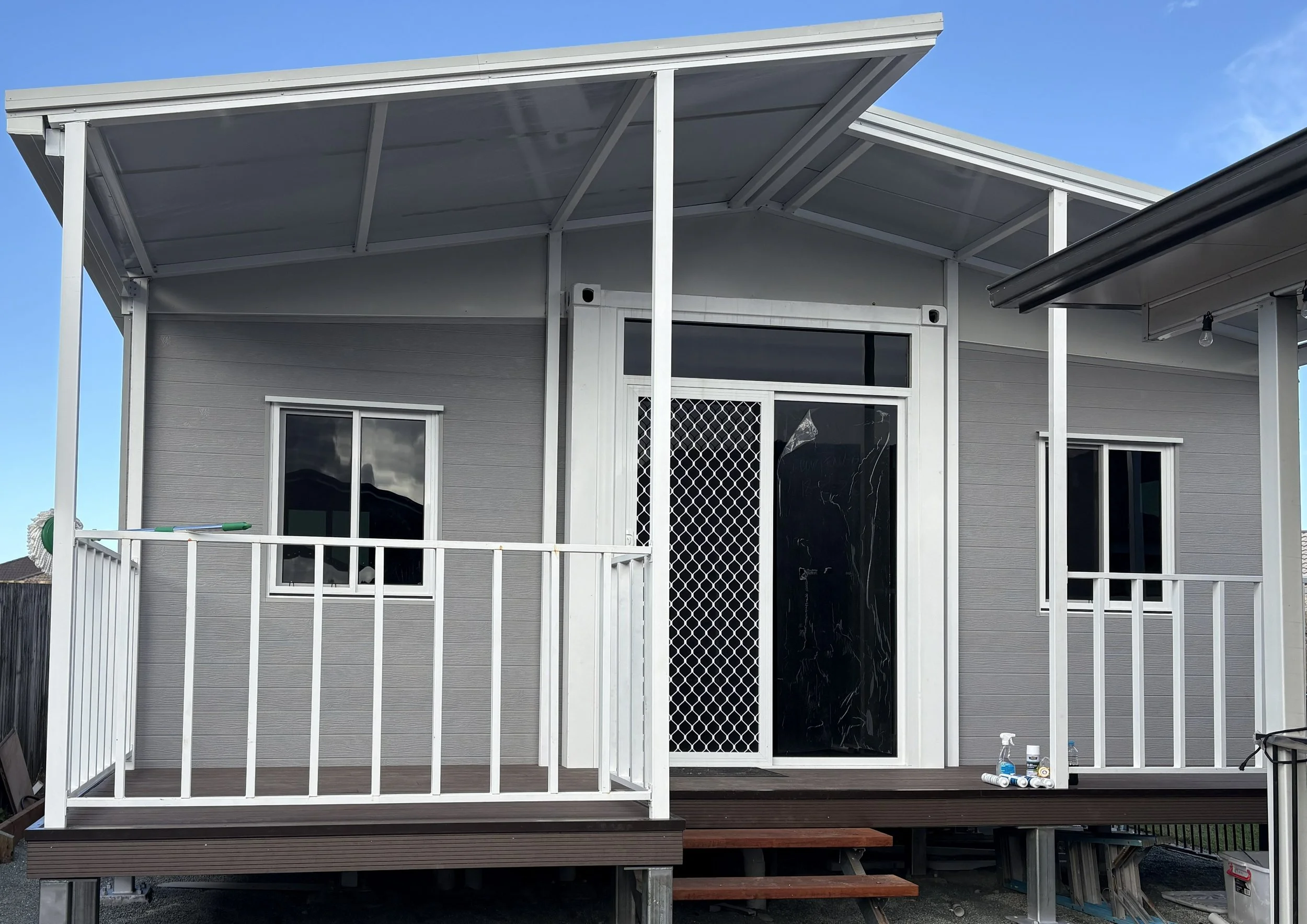 A small house with a deck and a metal roof extension, several windows, and a sliding glass door, with tools and supplies on the deck and a clear blue sky in the background.