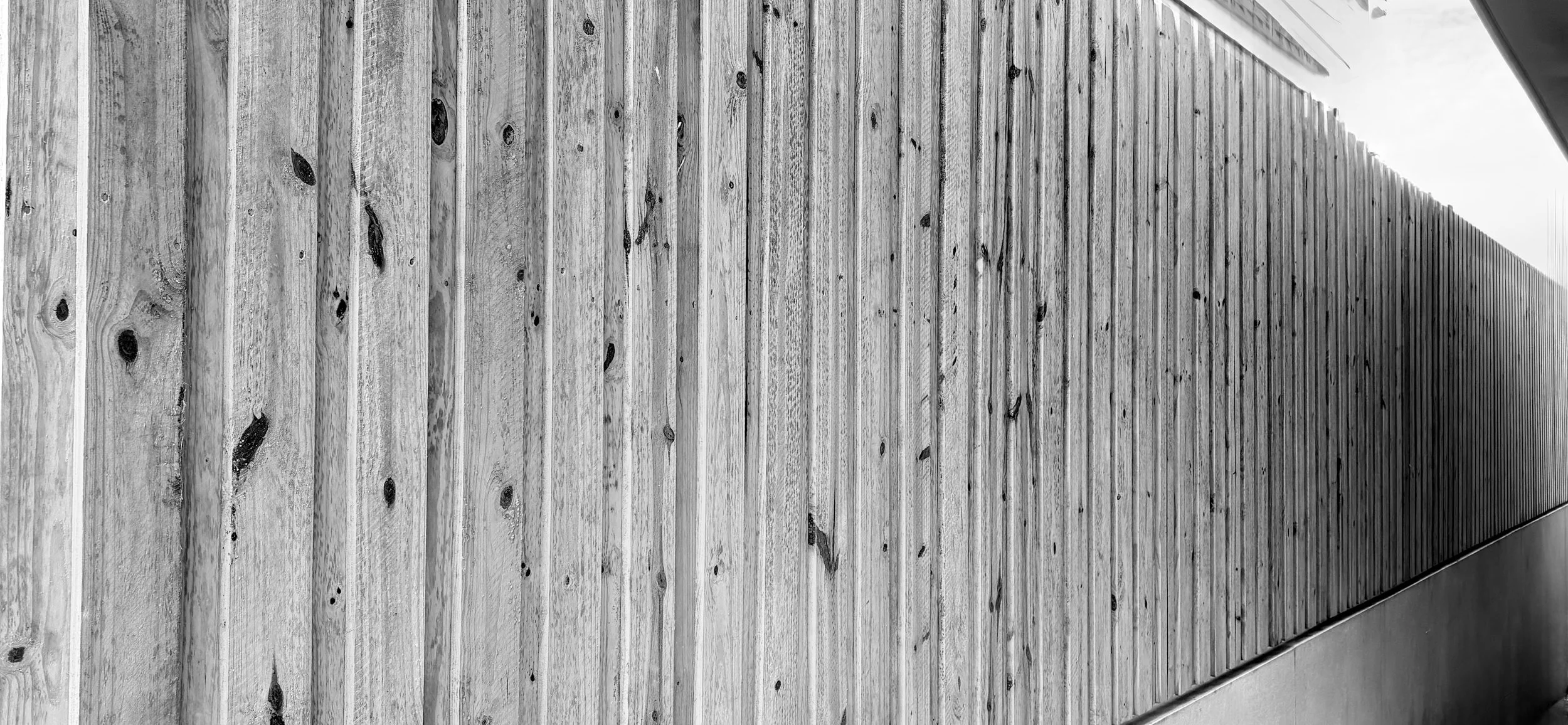 A long wooden fence made of vertical planks runs along a paved sidewalk, with a modern building wall partially visible to the right.