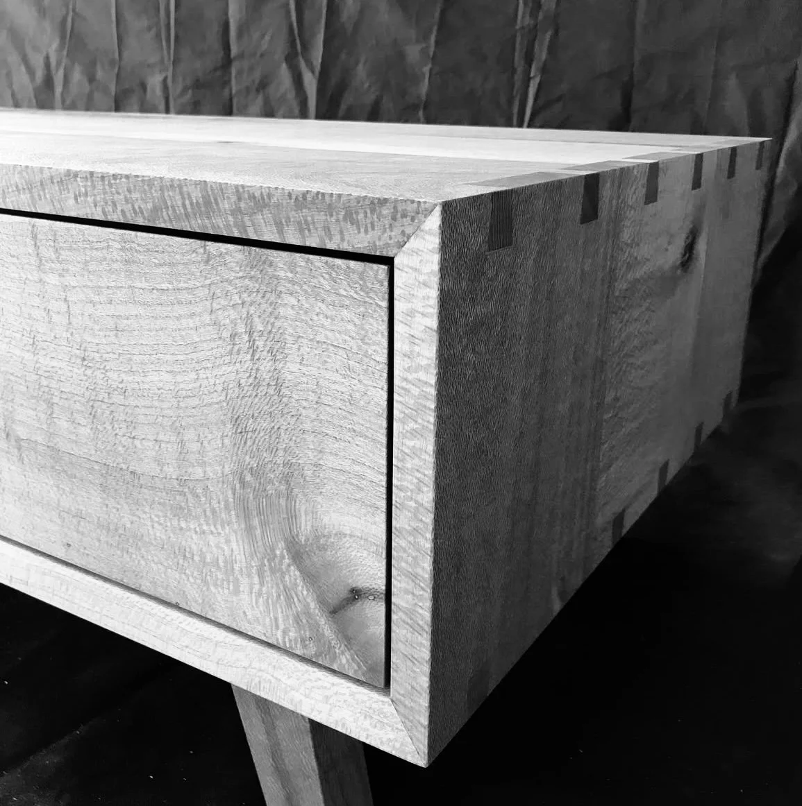 Close-up of a wooden piece with visible wood grain and dovetail joints, possibly part of a furniture piece, in black and white.