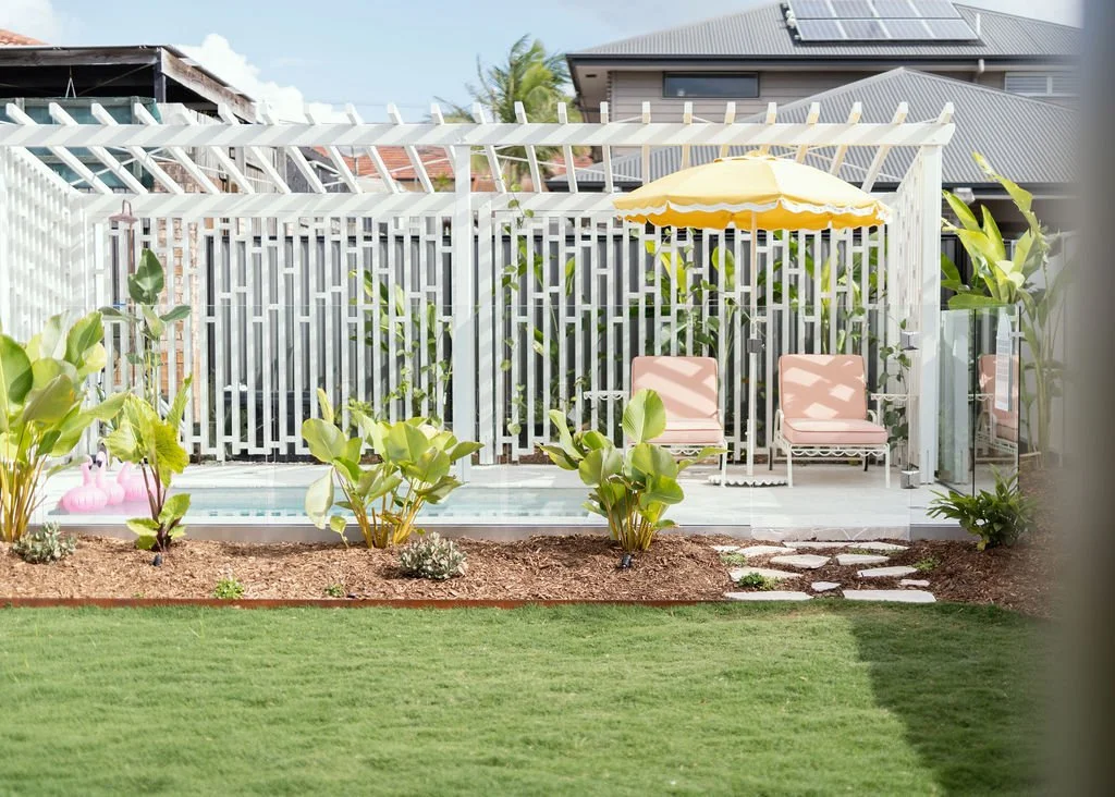 A backyard patio with two pink lounge chairs, a yellow umbrella, and a small swimming pool surrounded by plants and a wooden fence.