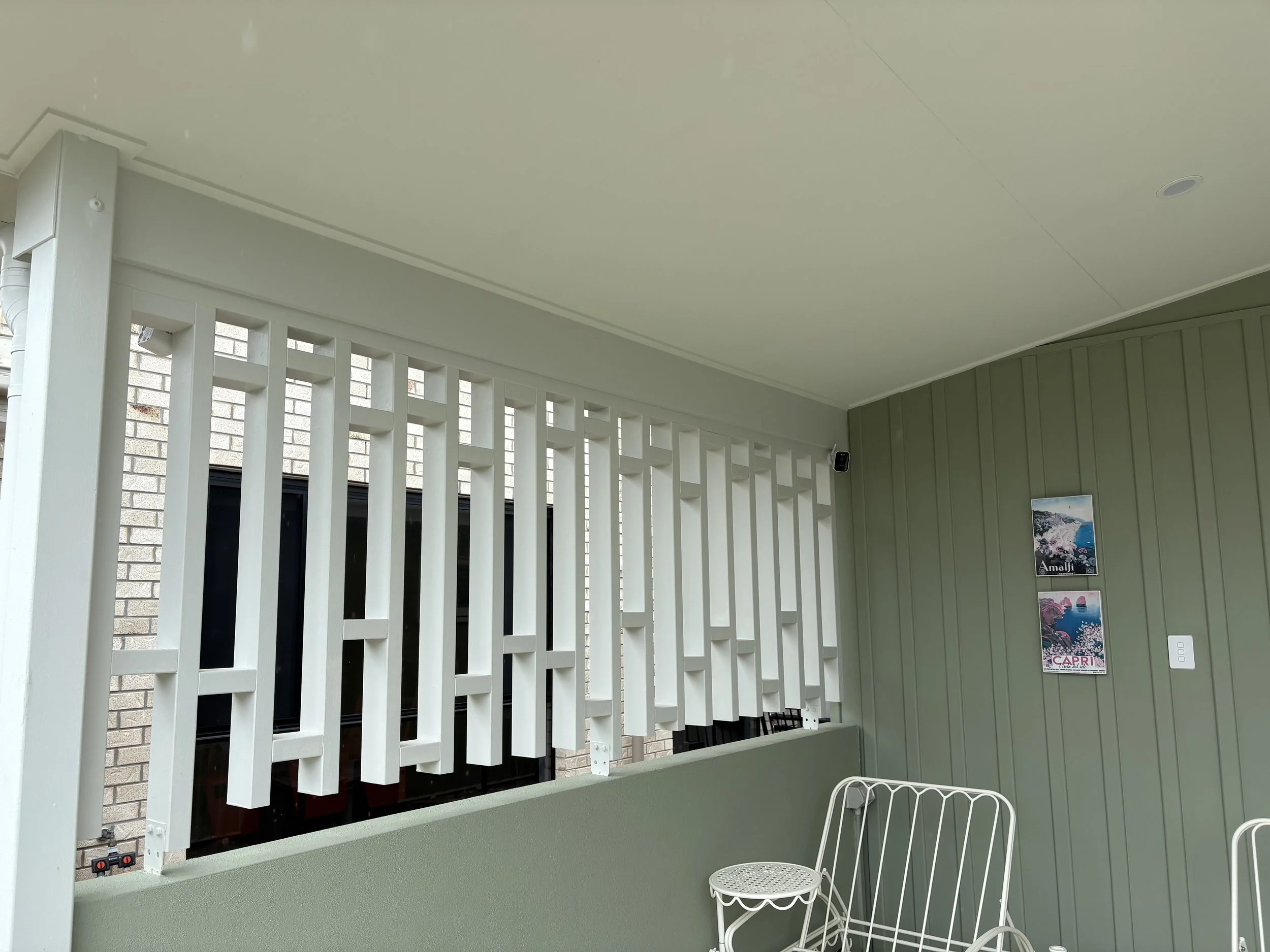 A covered outdoor patio area with green painted walls and a white wooden lattice privacy screen. There are two white metal chairs and a landscape poster on the wall.