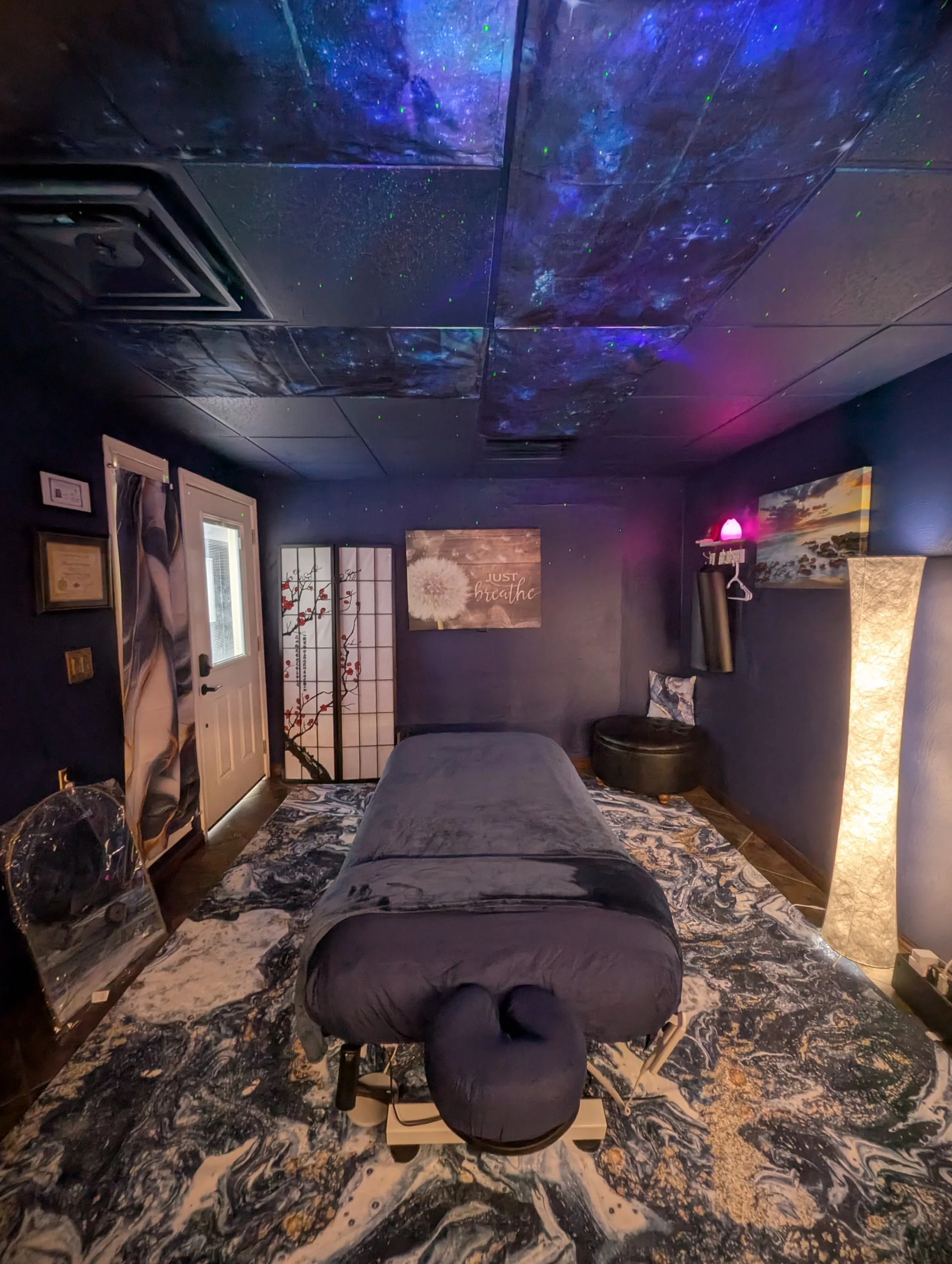 Picture of the professional massage room with a ceiling decorated to resemble outer space with stars and aurora lights, a massage table in the center, artwork on the walls, and a patterned rug.