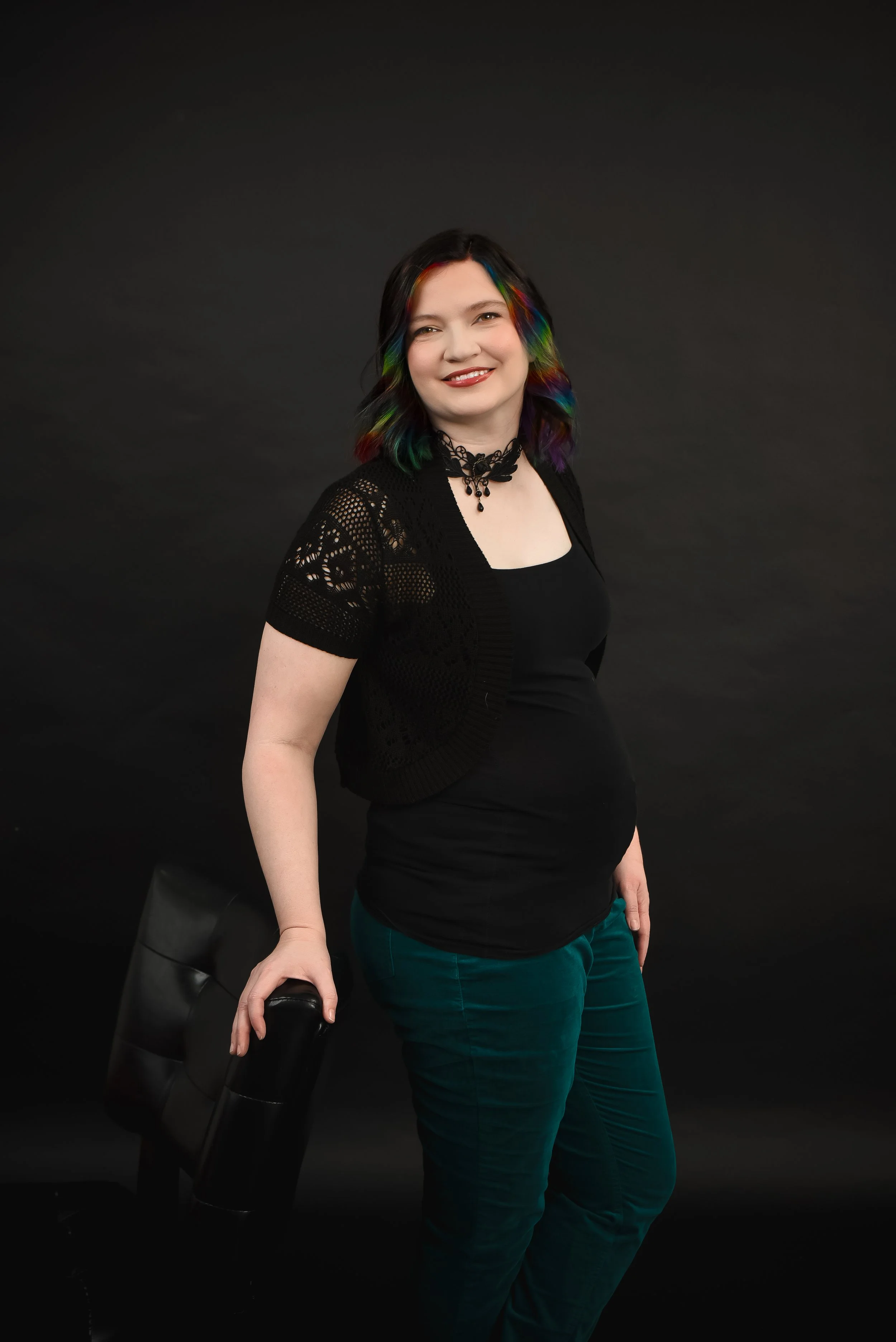 A woman with rainbow-colored hair, wearing a black top and a necklace, smiling while leaning on a chair against a black background.