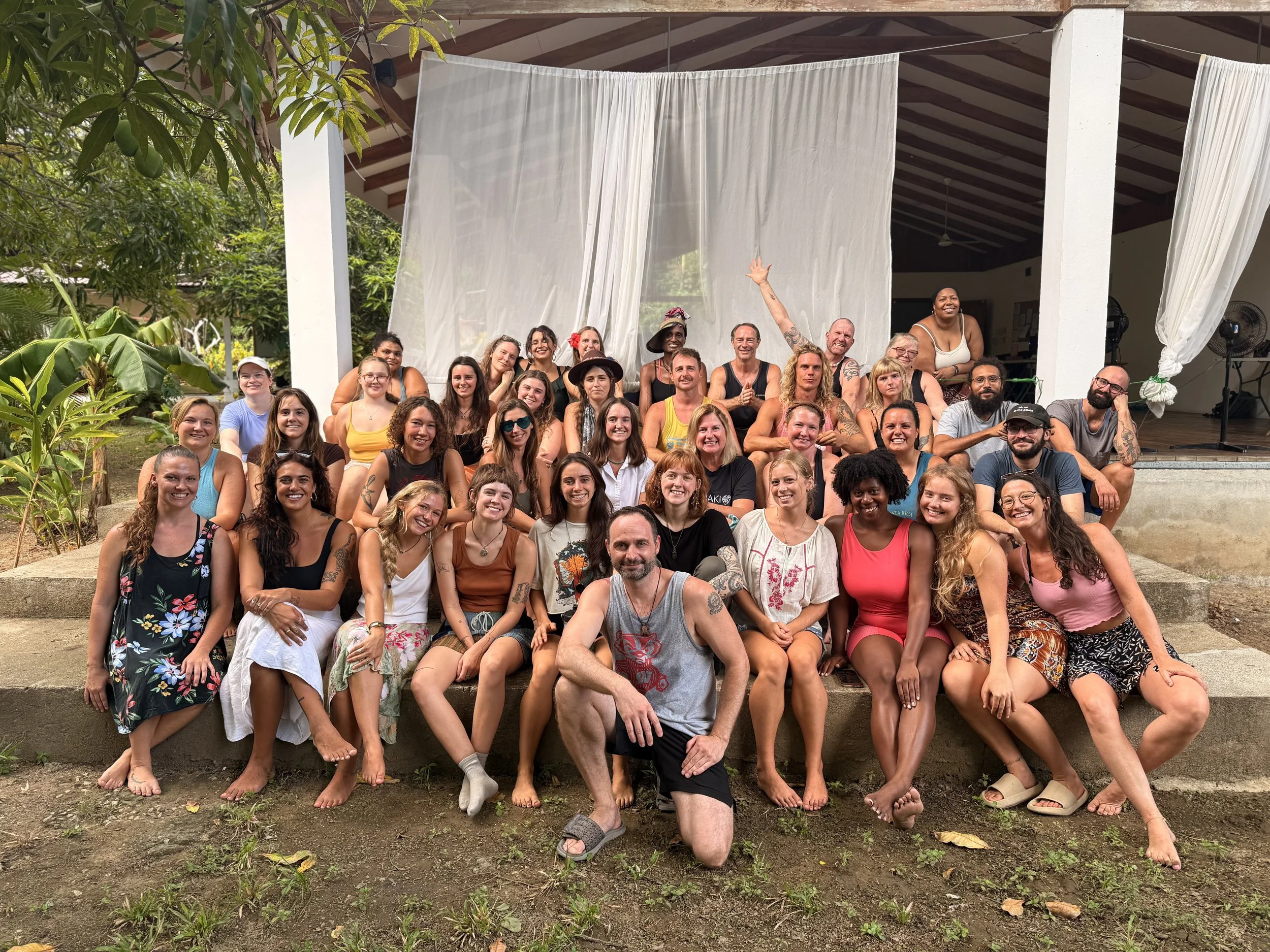 A large group of smiling people gathered outdoors on steps for a group photo, with a covered patio and greenery in the background.