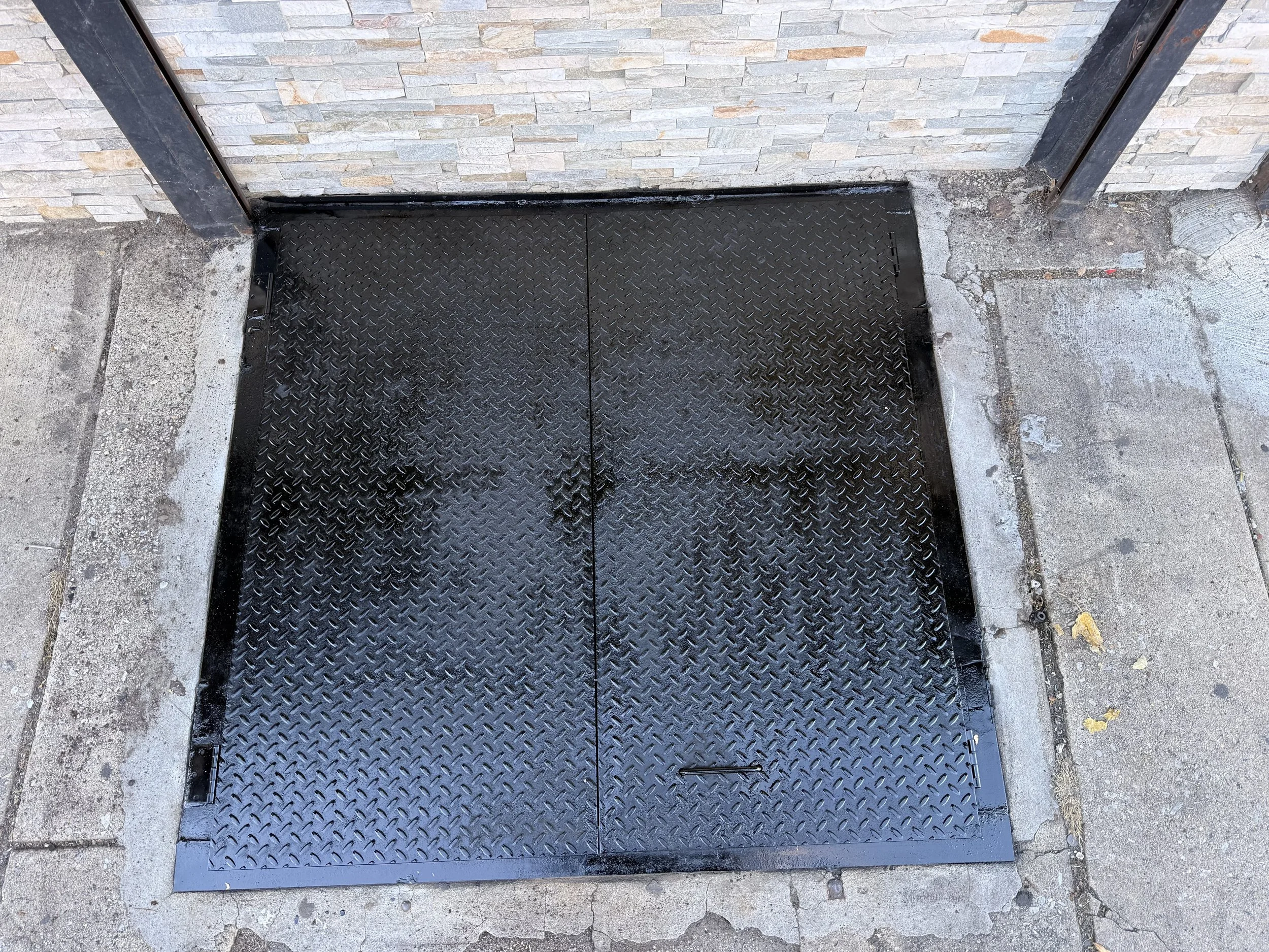 A black metal ramp with a diamond plate surface for wheelchair access, installed against a stone wall and concrete sidewalk.