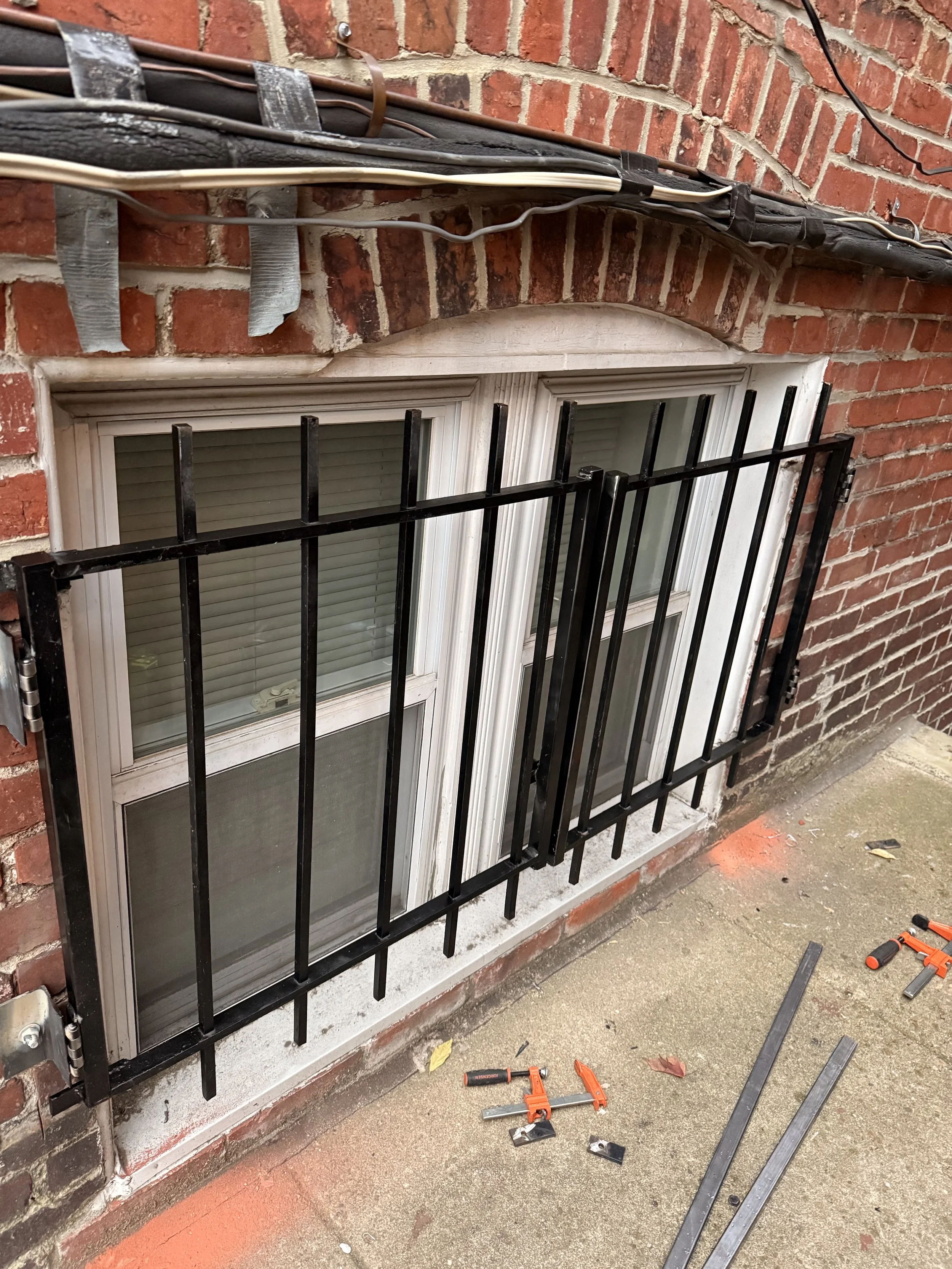 A window with black metal bars installed on a red brick wall. Tools and metal parts are on the ground nearby for installation.