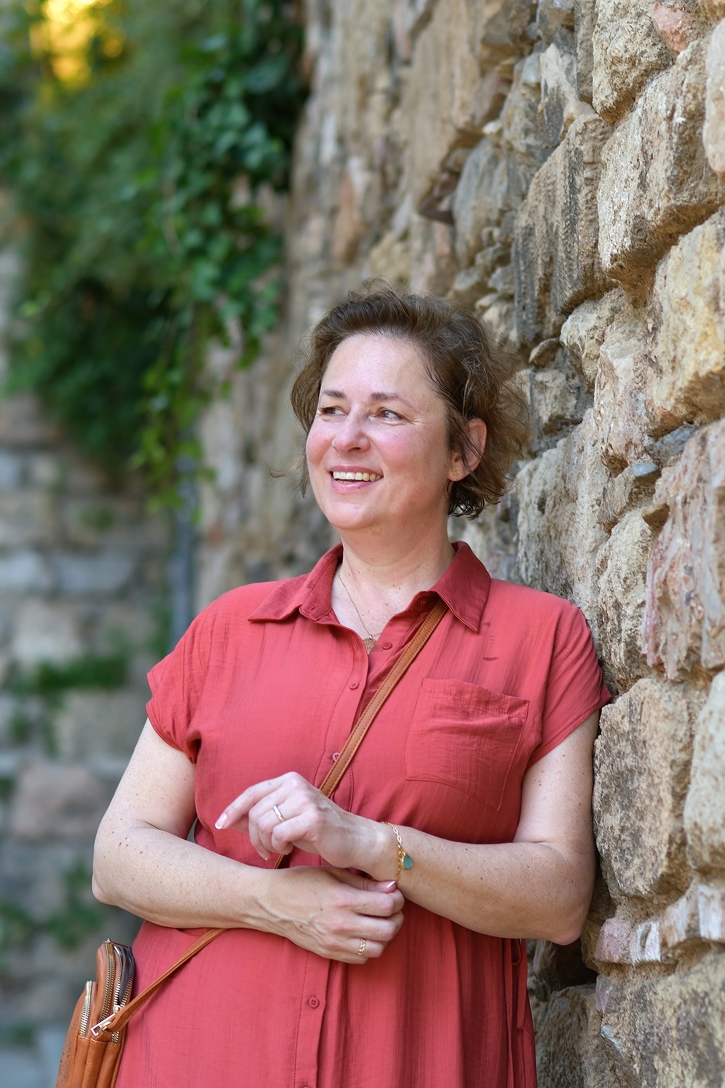 Marcela Kratochvilova smiling against a stone wall with ivy, wearing a coral shirt