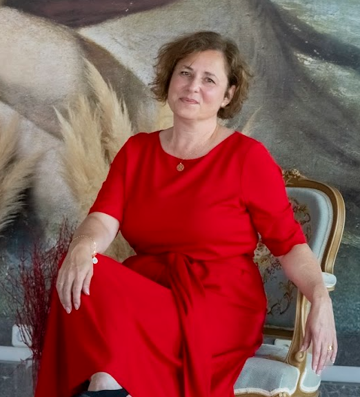 Marcela Kratochvilova seated in a wooden chair wearing a red dress, composed and thoughtful expression