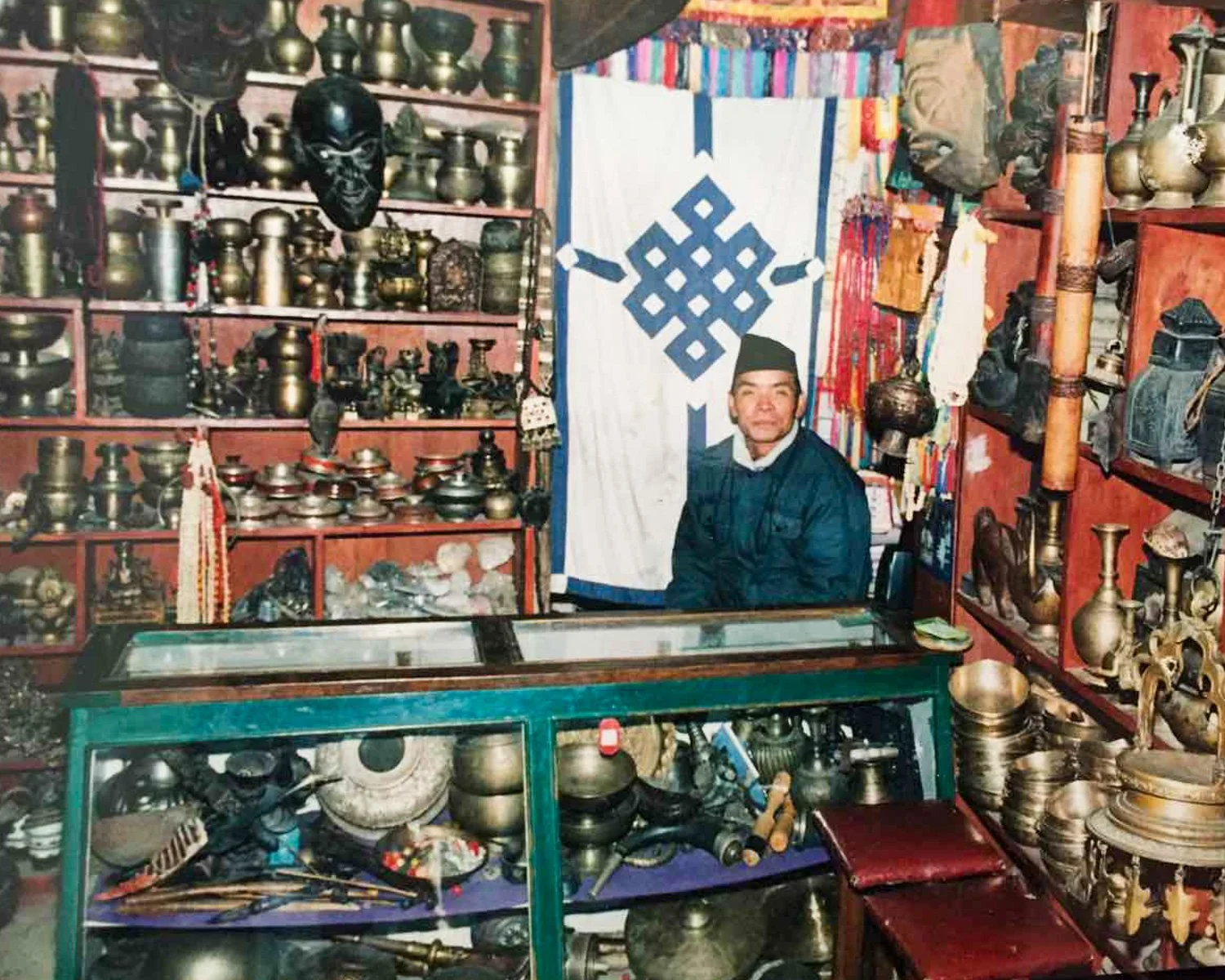 A man sitting inside a shop filled with brass, bronze, and metal artifacts and jewelry, with shelves of various decorative items and a cloth with a blue geometric pattern hanging on the wall behind him.
