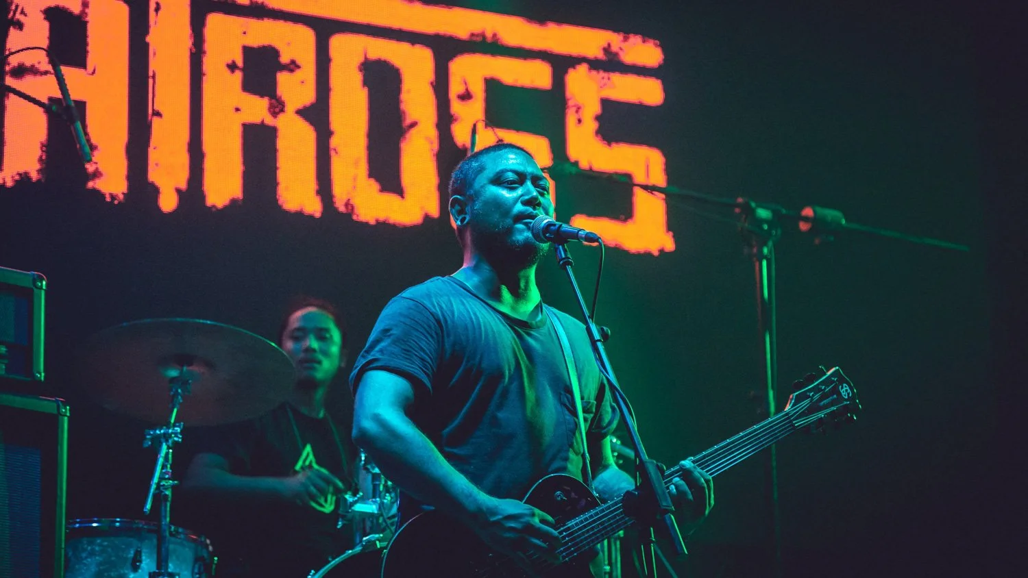 Musician playing an electric guitar and singing into a microphone on stage during a concert, with a drummer in the background and a large illuminated sign behind them.