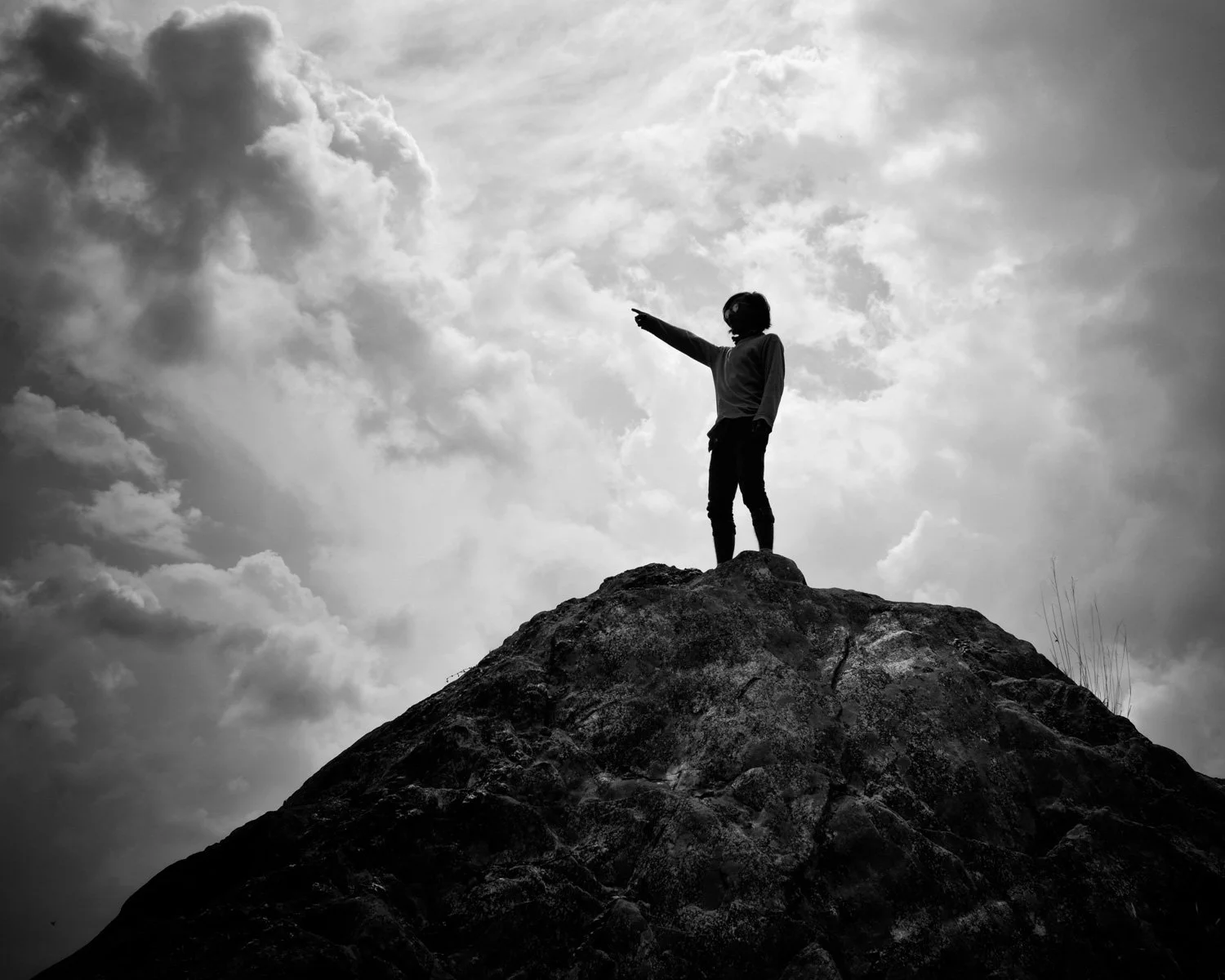 Silhouette of a person standing on a large rock, pointing into the distance, against a cloudy sky in black and white.