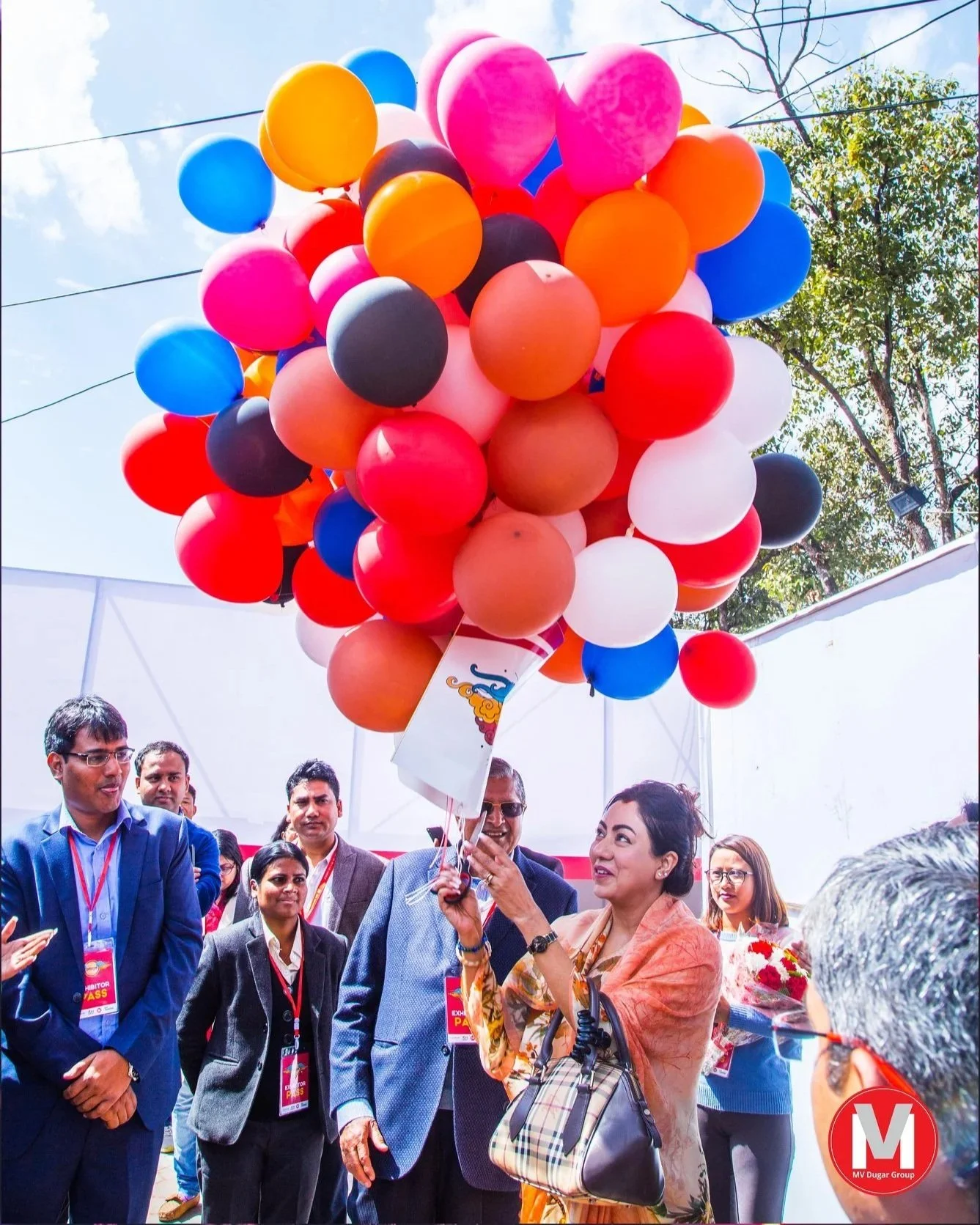A group of people, including a woman in an orange sari, releasing colorful balloons at an outdoor event. The balloons are various colors including pink, orange, blue, red, and black, with some tied with strings. The background features a partly cloud
