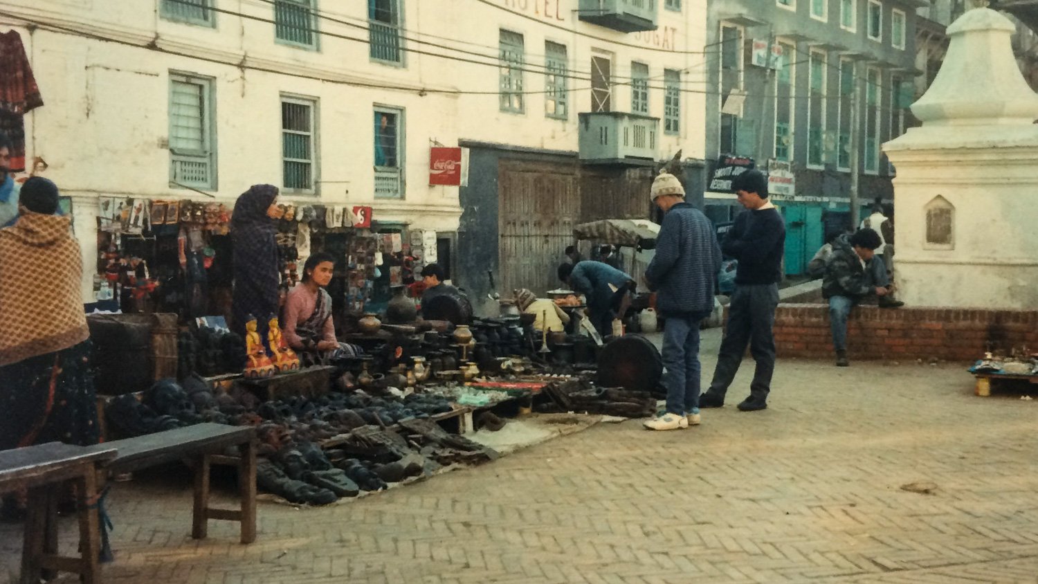 Street market with various vendors selling shoes, pottery, and accessories among people walking and shopping, with buildings in the background.