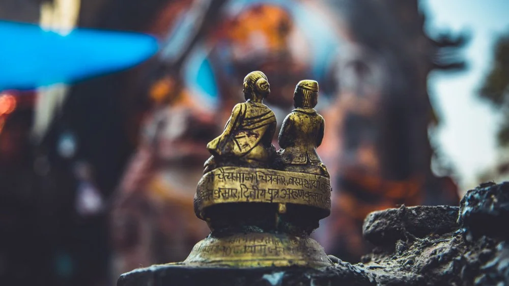 A small brass figurine of two people sitting on a platform, with inscriptions, placed on a rocky surface, with a blurred colorful background.