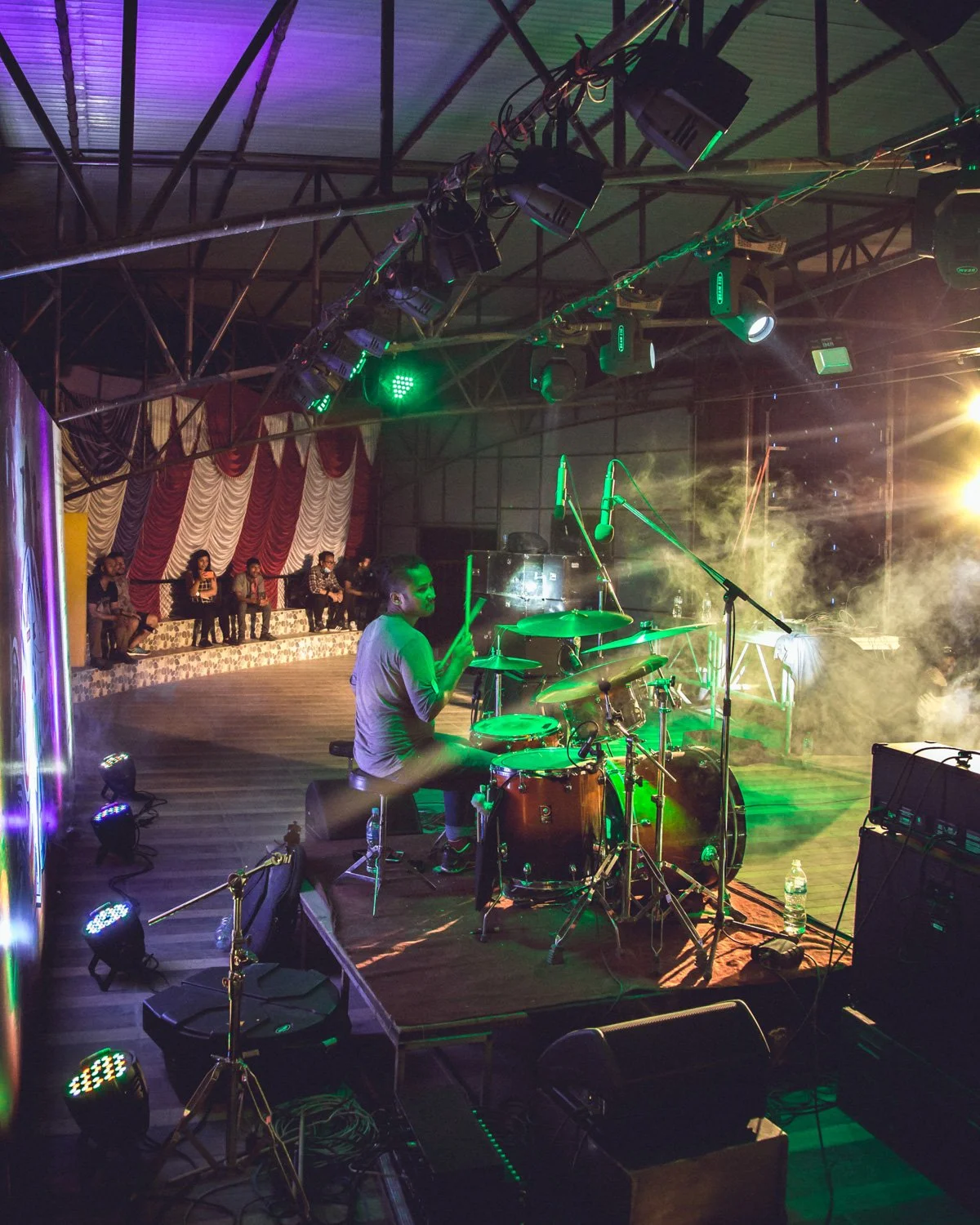 A musician playing the drums on stage with green lighting, smokey atmosphere, and an audience sitting on a raised platform in the background.