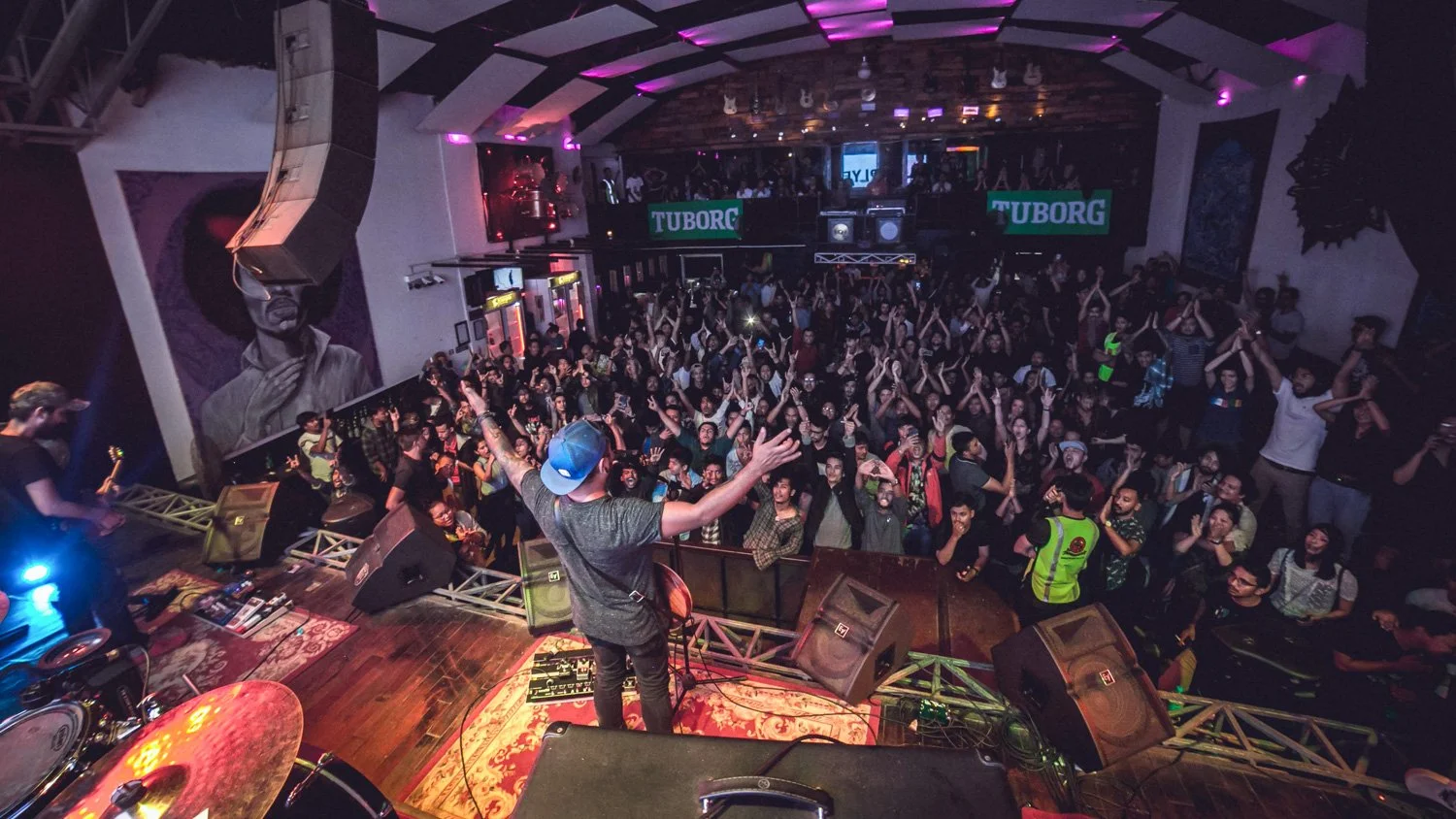 Crowd at a nightclub dancing and having fun with a DJ performing on stage. The venue is decorated with posters and neon signs, including two green Tuborg signs. People are cheering and enjoying the music.