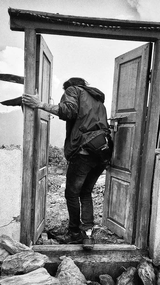Person with tattoos and dark clothing opening wooden gate outdoors, rocks and cloudy sky visible