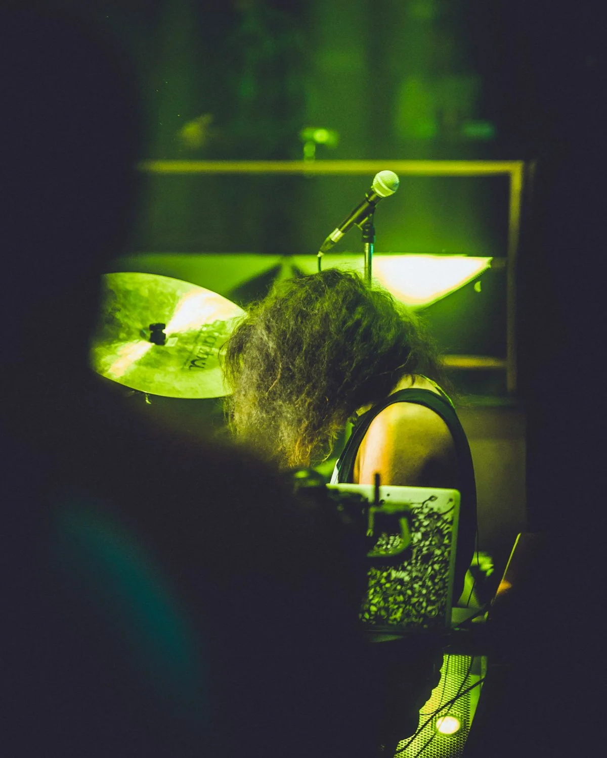 A person with curly hair sitting at a drum kit during a live music performance with green stage lighting, with a microphone above them and a cymbal in the foreground.