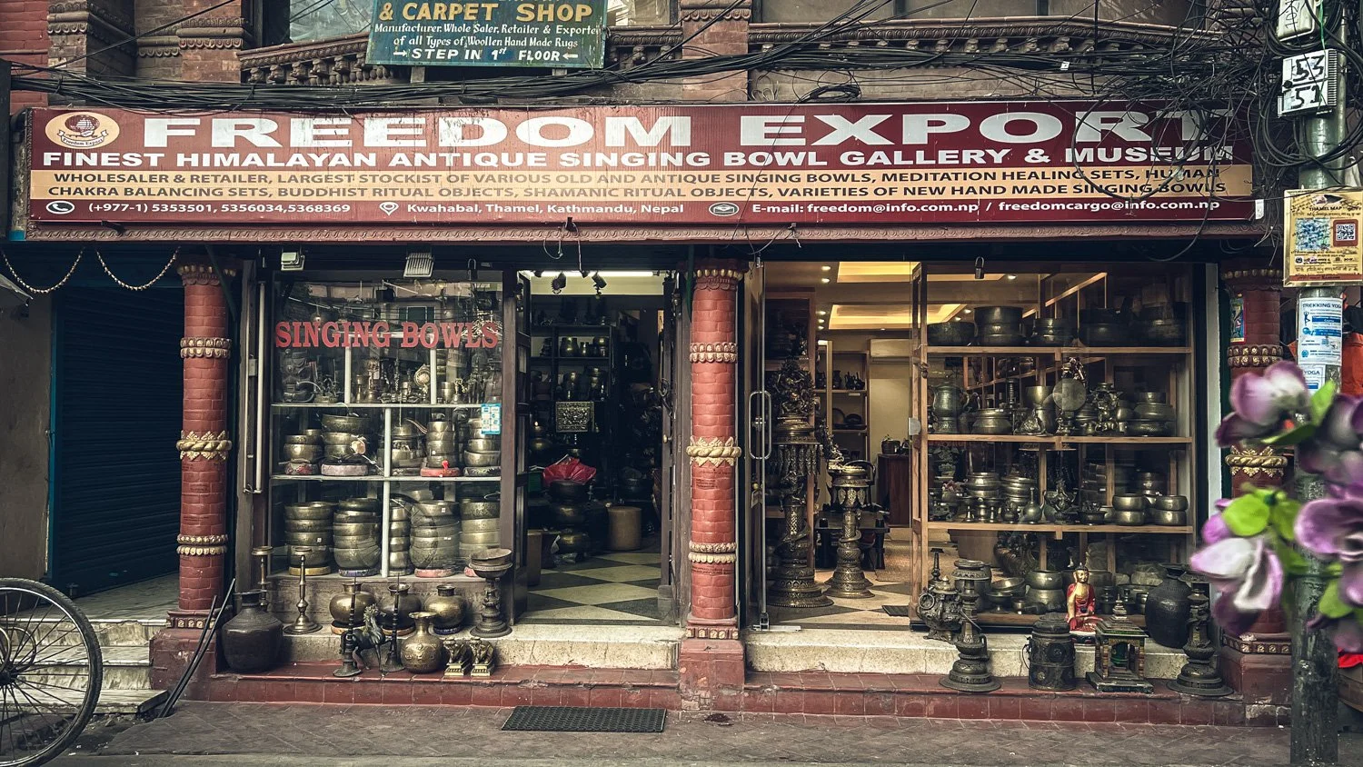A shop in Kathmandu, Nepal, named Freedom Export, selling antique singing bowls and related items. The shop has shelves filled with metallic singing bowls, and the storefront displays various antique metalware, statues, and artifacts.
