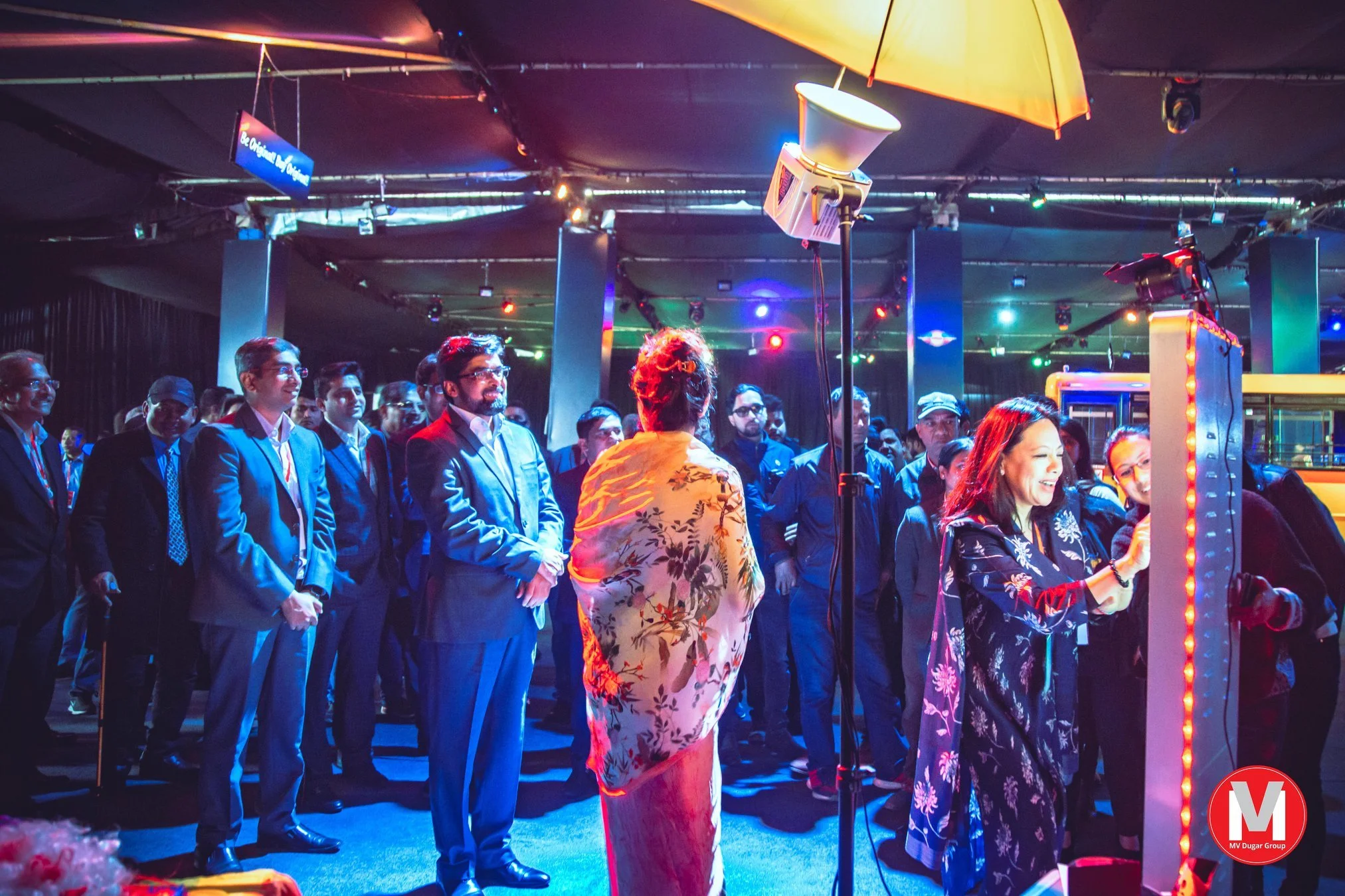 People gathered indoors at an event, with some wearing formal suits and dresses. There are colorful lights and equipment, including a yellow umbrella over a stage area, and a woman in a floral dress speaking to the crowd. The atmosphere appears livel