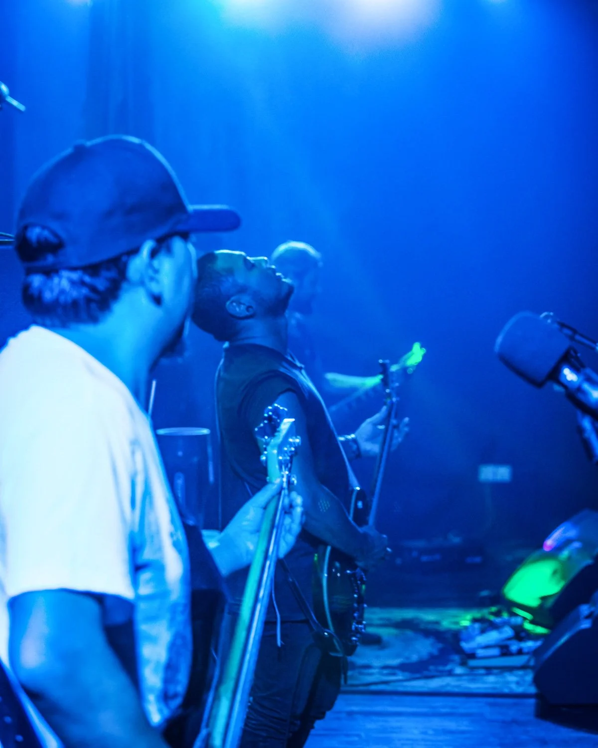 Three people standing in a dark room with blue lighting, holding microphones and musical instruments, possibly performing or rehearsing.