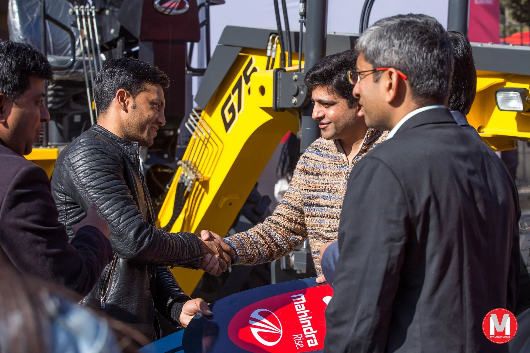 Two men shake hands during an outdoor event, with a yellow construction vehicle in the background. Several other people are present, and one is holding a sign with the VN logo and red text.