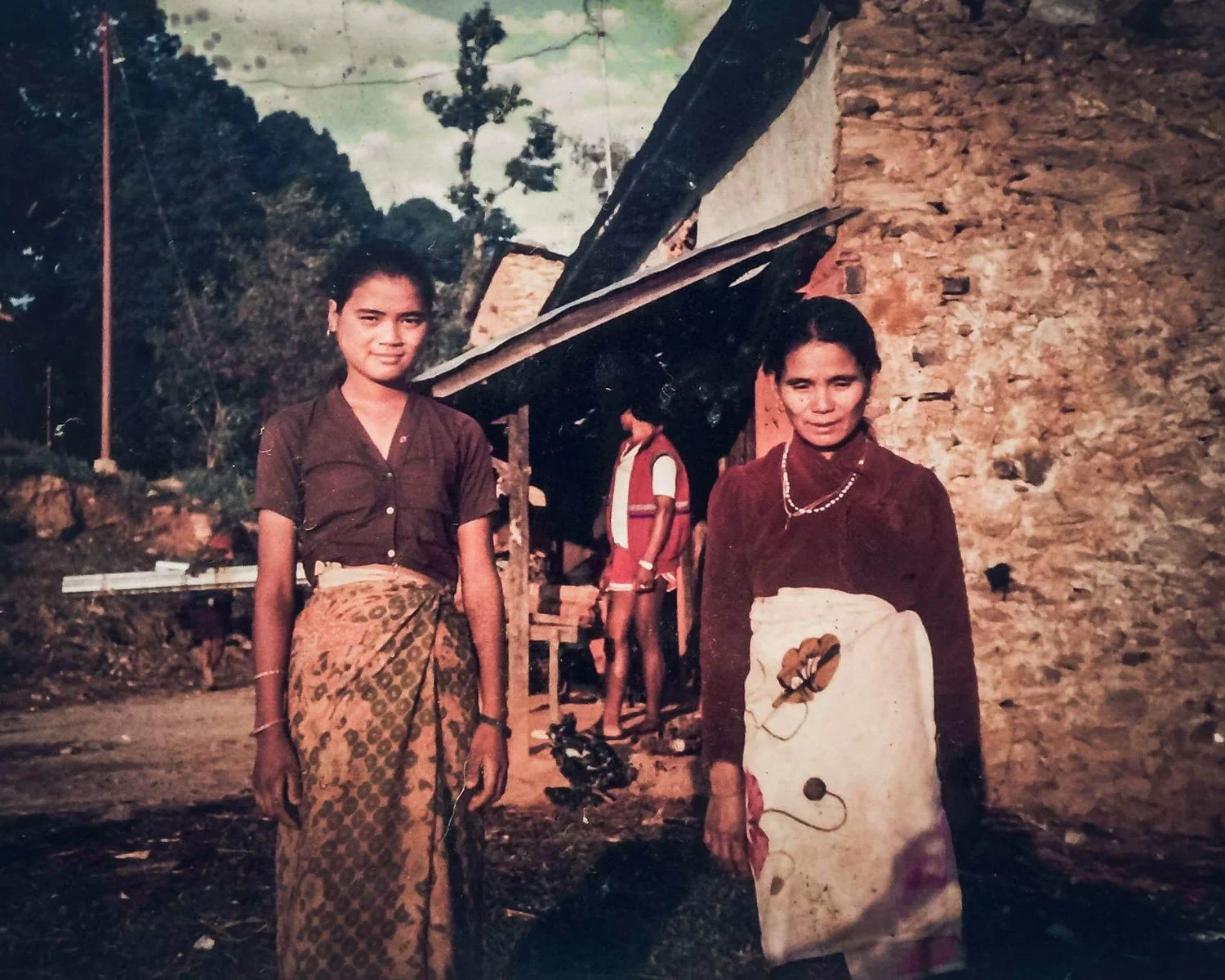 Two women standing outdoors in front of a rustic stone building, one with a patterned skirt and the other with a beige apron, with a child in the background near the building.