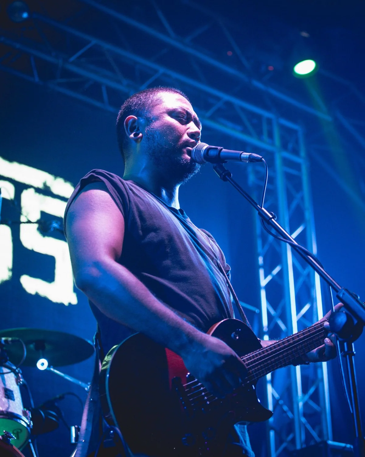A male musician singing into a microphone while playing an electric guitar on stage, with colorful stage lights and metal trusses in the background.