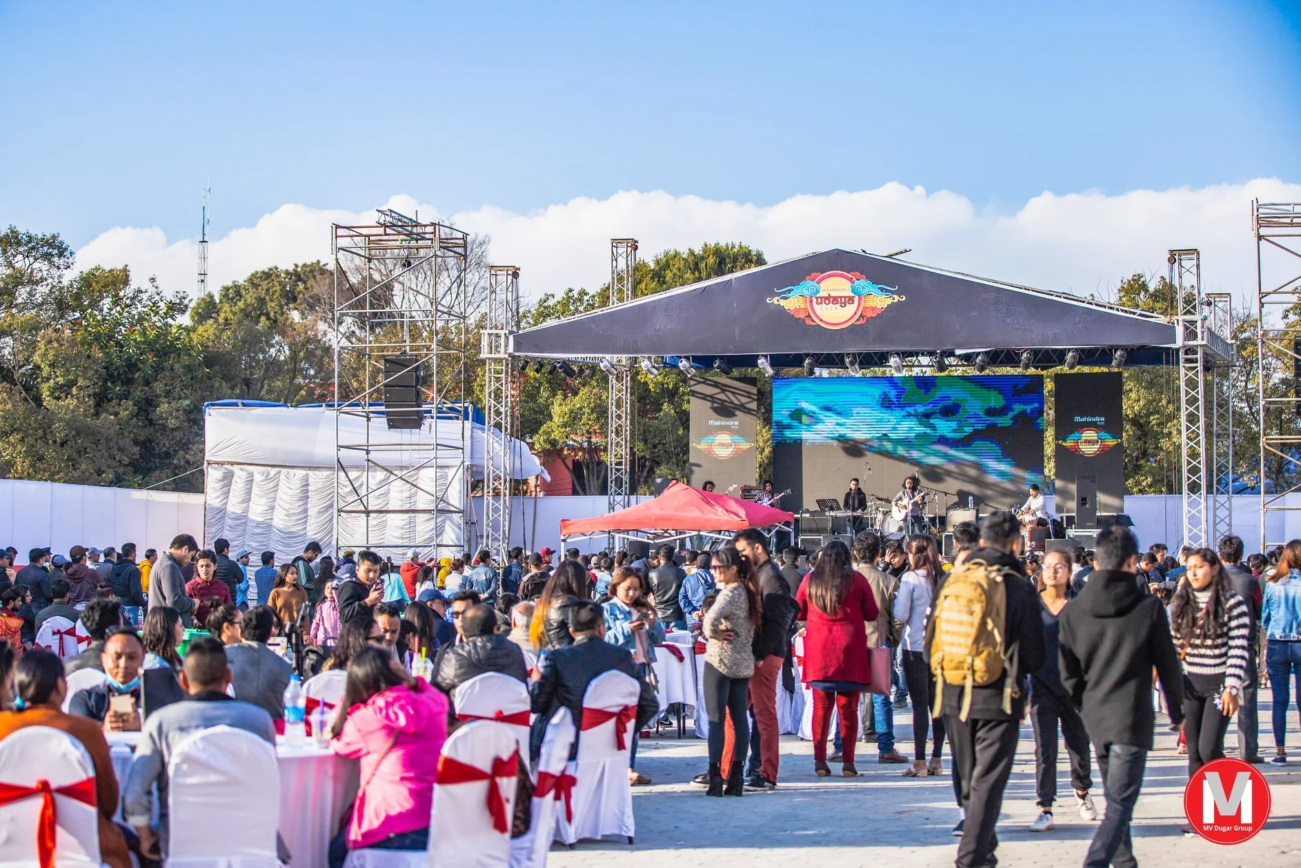 A large outdoor concert during daytime with a crowd of people standing and sitting, facing a stage with performers playing musical instruments. The stage has a large digital screen and is decorated with a banner that reads "Udaya." Trees and clear bl