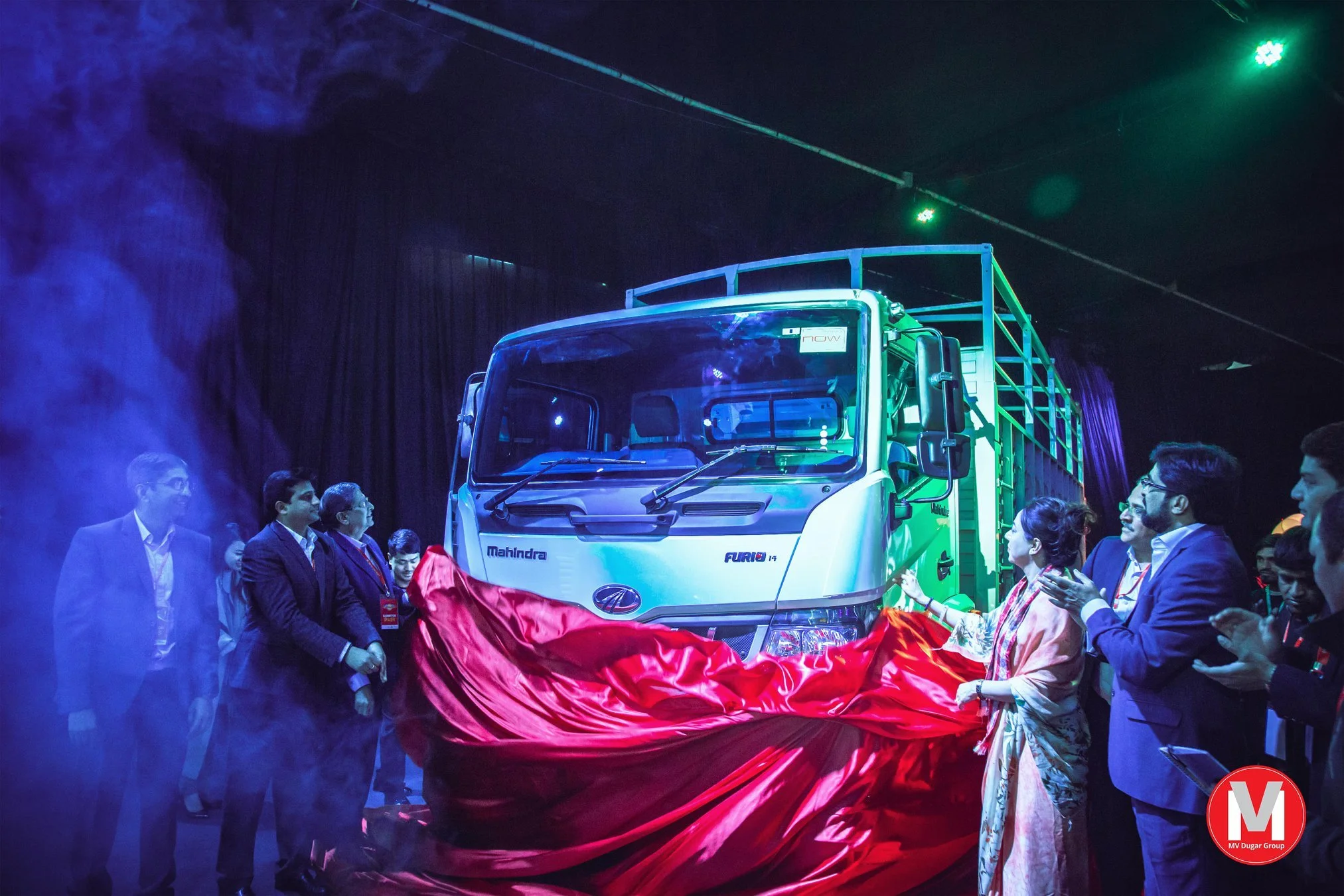 A group of people, dressed in business and traditional attire, gathered around a new green and white Mahindra FURIO truck during an unveiling event, with a red cloth partially covering the vehicle. The scene is illuminated with blue and green lightin