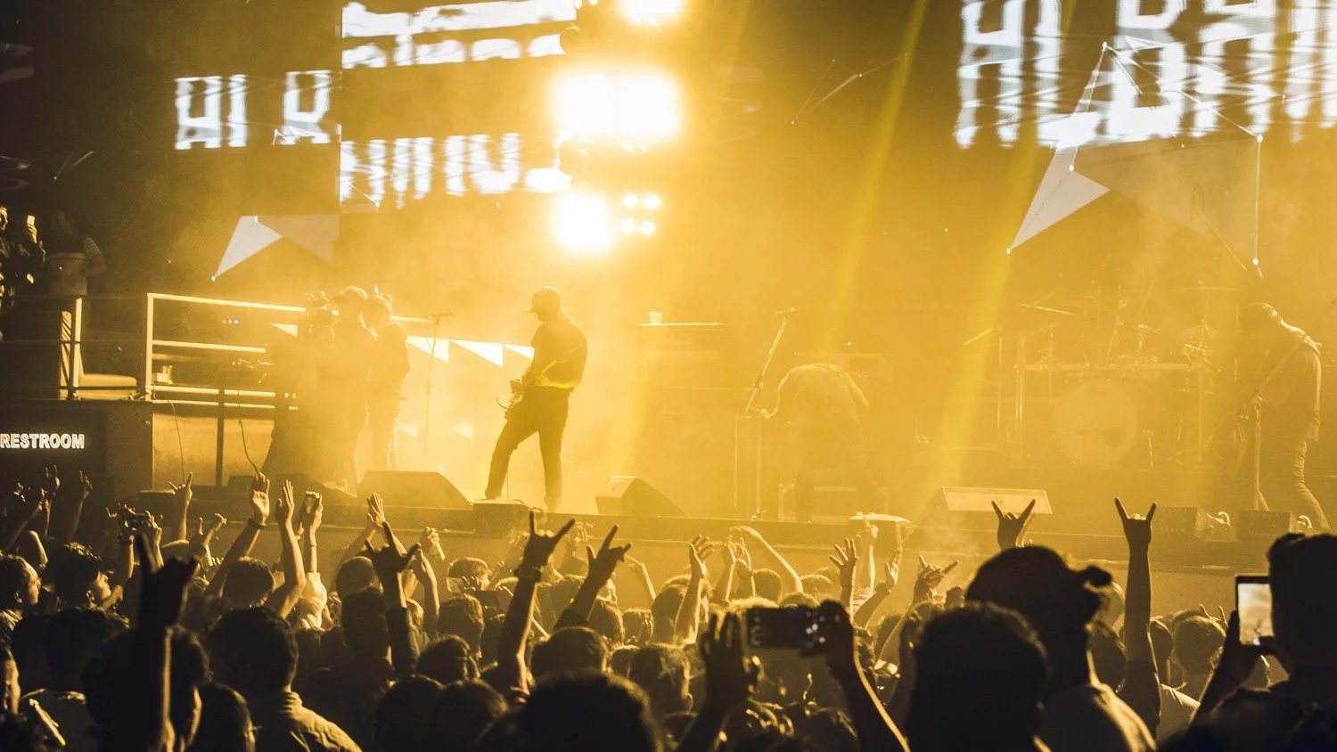 Concert scene with a band performing on stage illuminated by yellow lights, crowd seen with hands up, some taking photos.