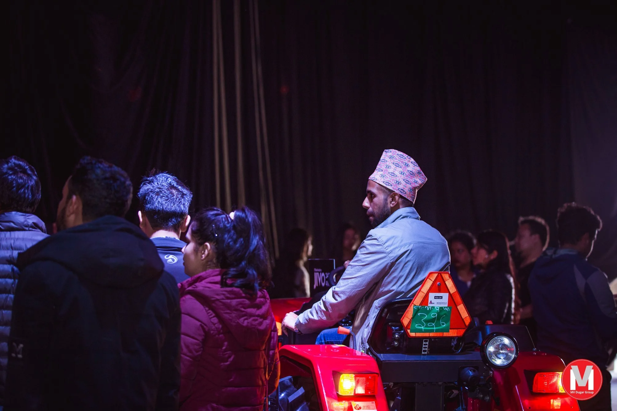 People gather in a dimly lit indoor event space, with a man in a patterned hat sitting in a mobility scooter in the foreground. The scene includes various attendees, some wearing jackets, with dark curtains or drapes in the background and a red and g
